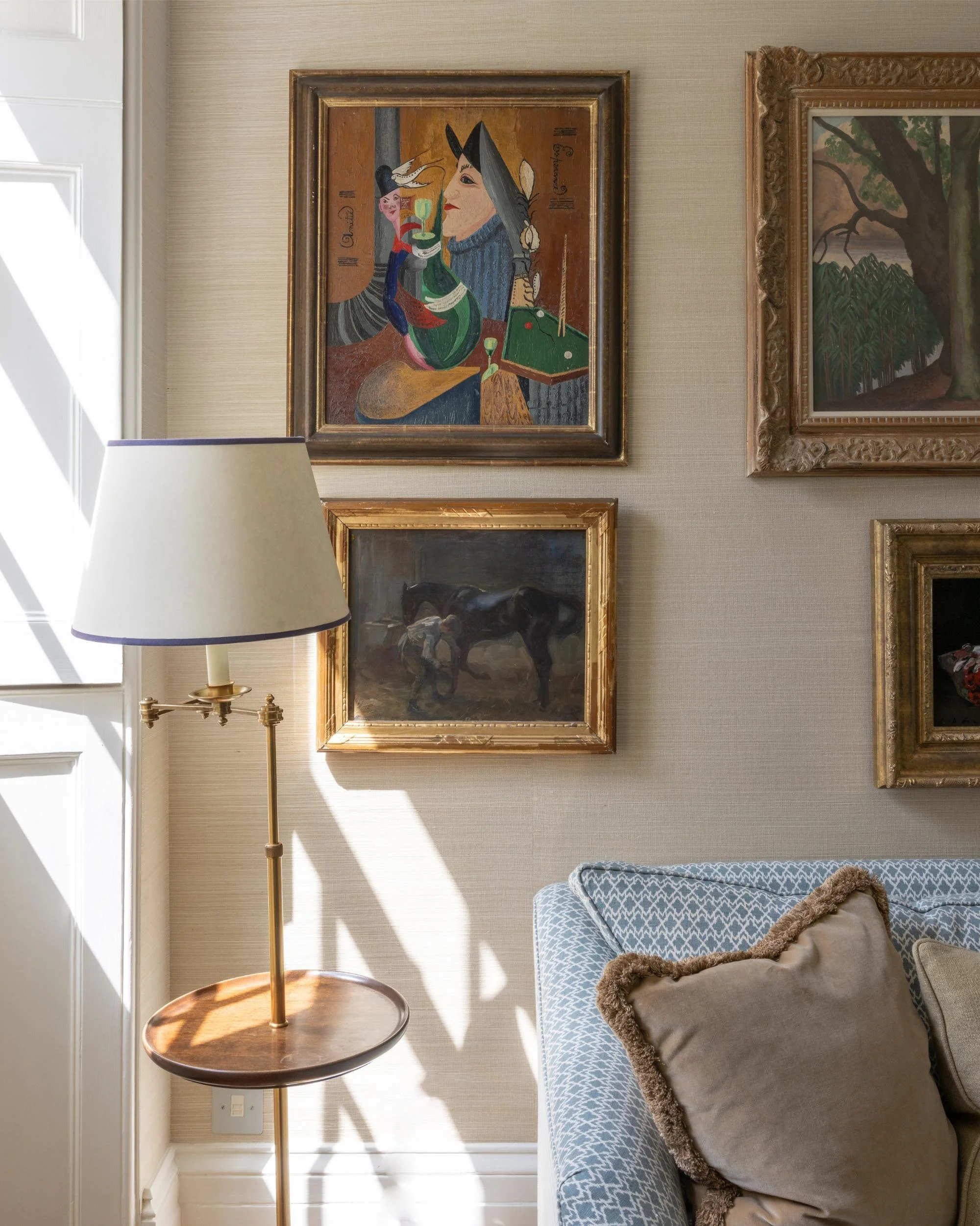 Interior scene of a living room wall with three framed paintings, a floor lamp with a beige shade, and a couch with pillows. The paintings include a colorful abstract portrait, a landscape with a large tree, and a dog.