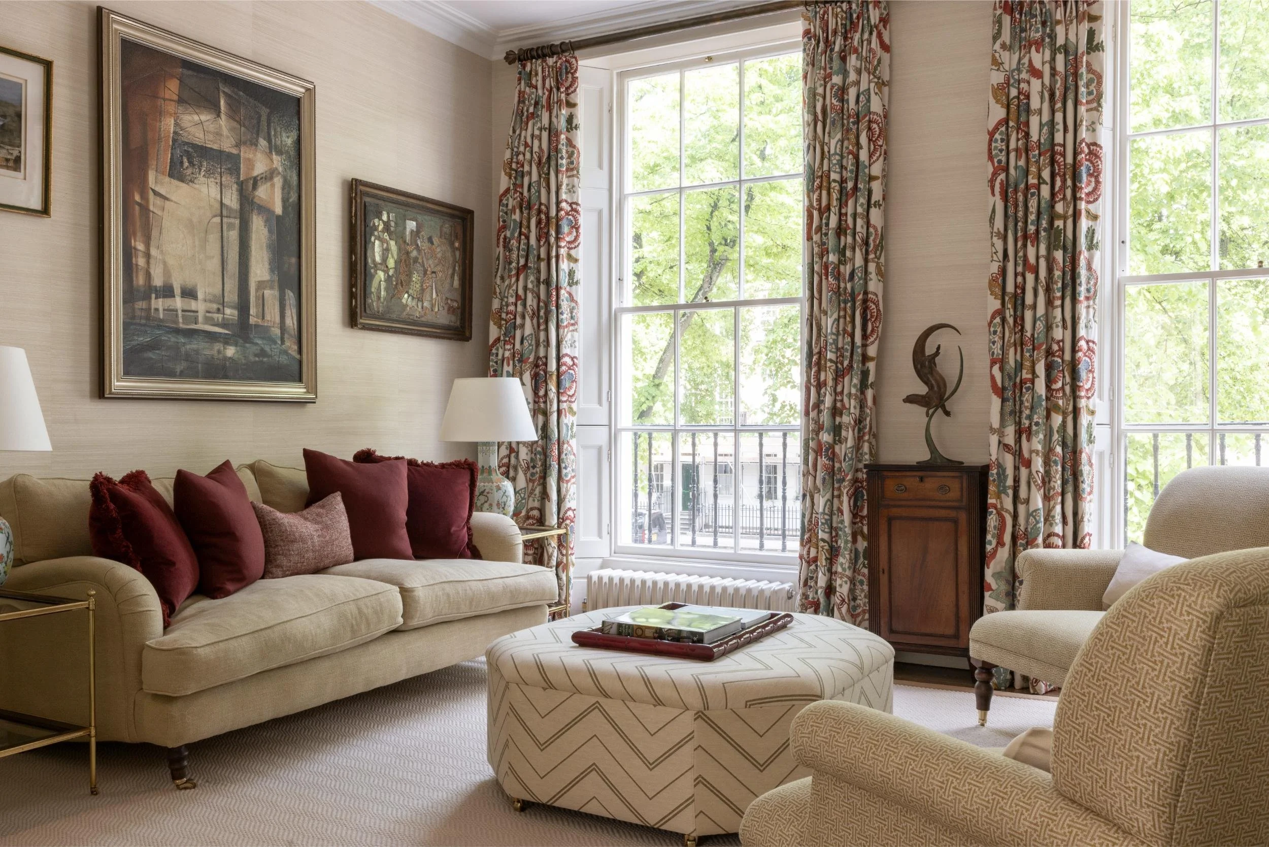Living room with cream-colored sofa, maroon and beige throw pillows, patterned armchairs, large window with floral curtains, framed artwork on walls, a wooden side table with a sculpture, and a round ottoman with books.