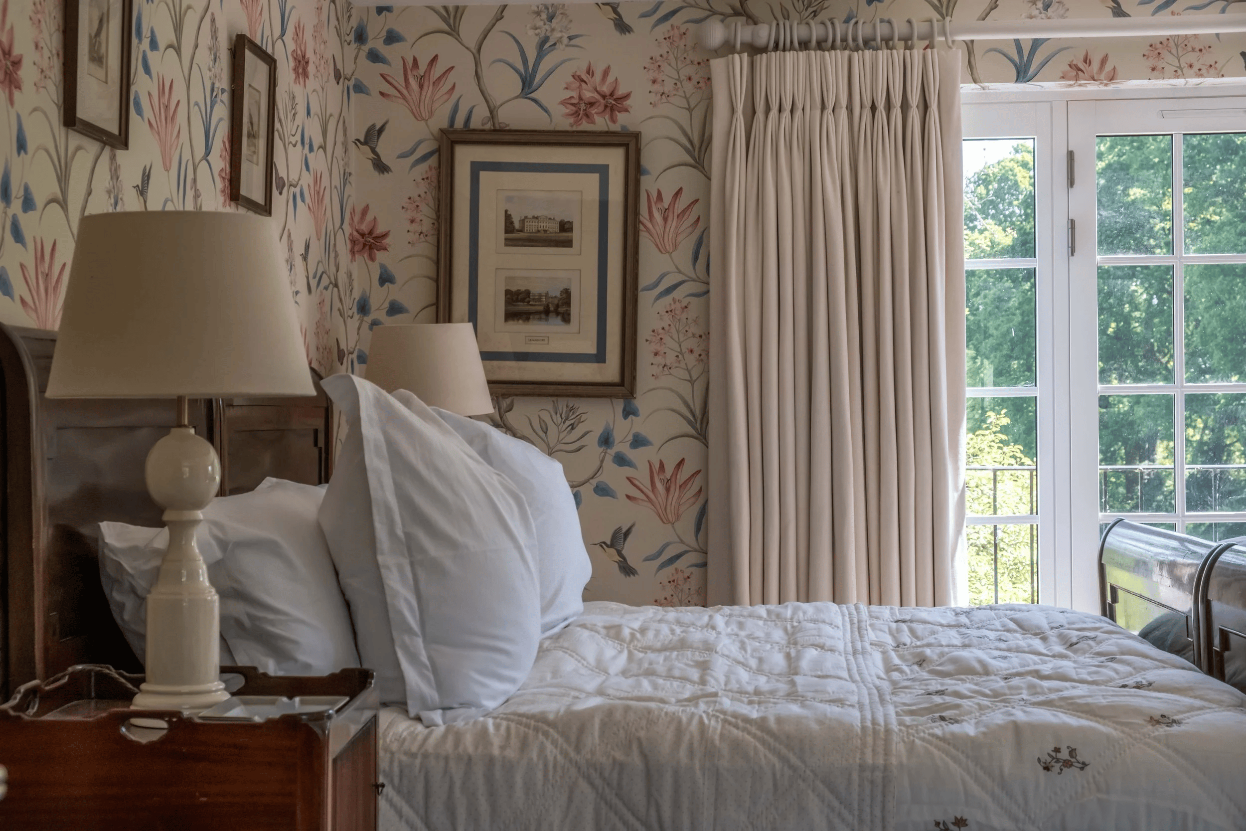 A bedroom with a neatly made bed featuring white pillow and bedding. There are two table lamps on wooden nightstands on either side of the bed. The wall behind the bed has floral wallpaper with pink, blue, and gray flowers and hummingbird illustratio