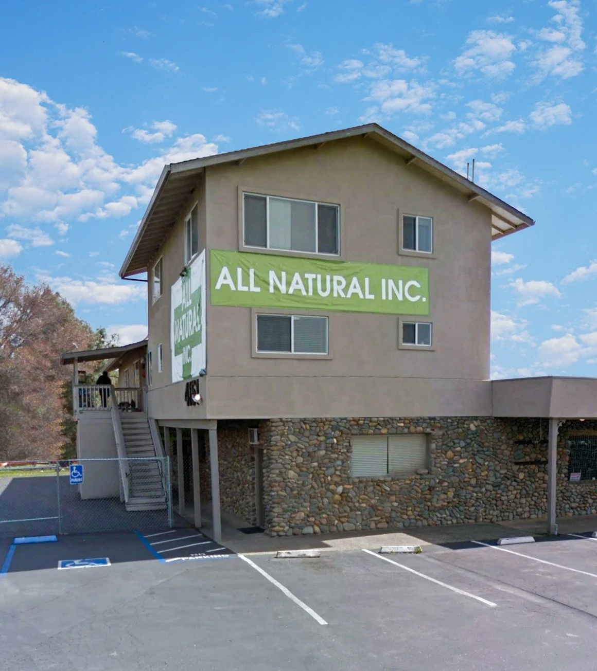 A multi-story building with a large green banner that reads 'ALL NATURAL INC.' The building has beige walls, multiple windows, and a stone facade on the lower level. There are parking spaces, including handicap designated spots, in the foreground.