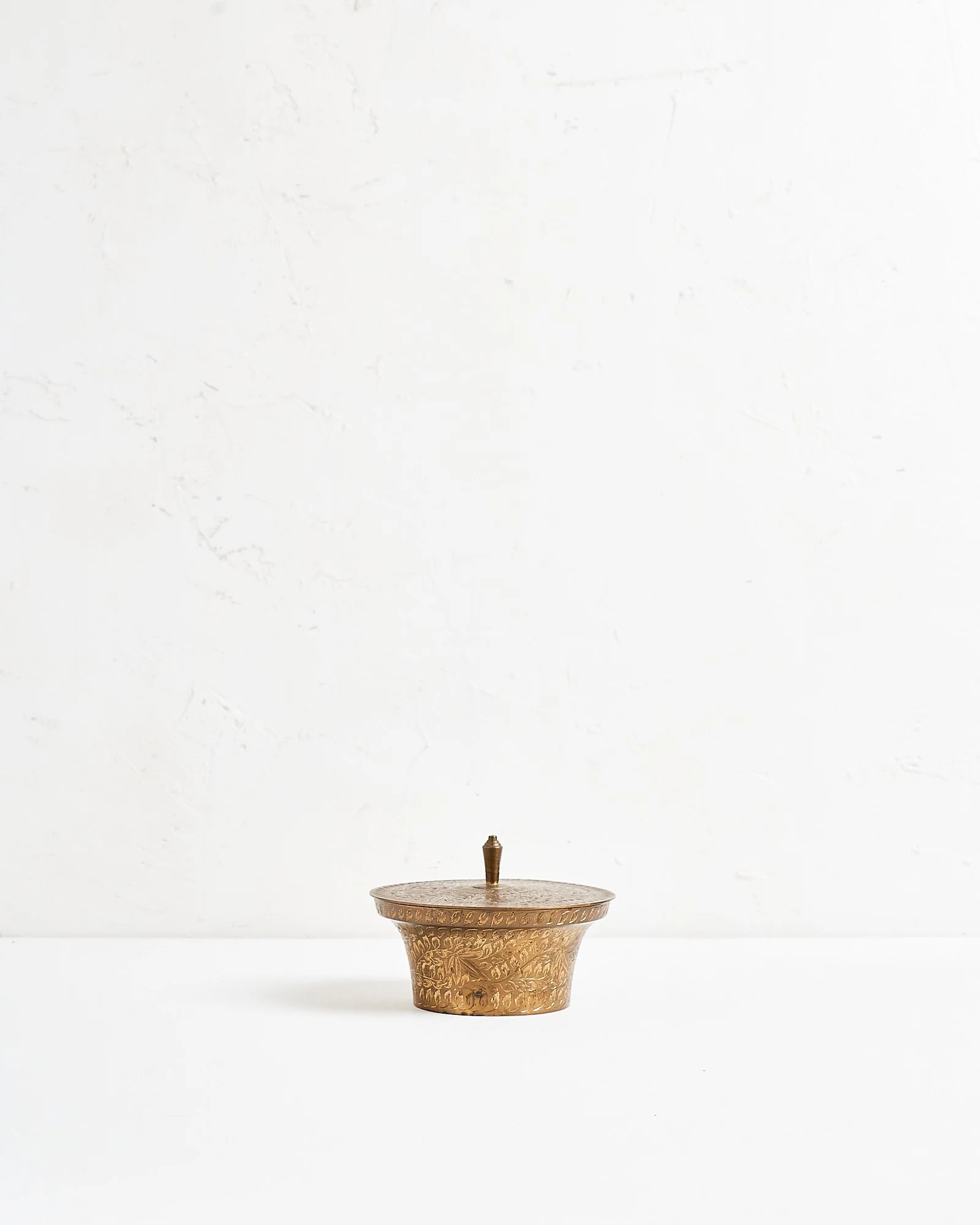 A round small brass decorative box with engraved patterns and a lid, sitting on a white surface against a plain white wall background.