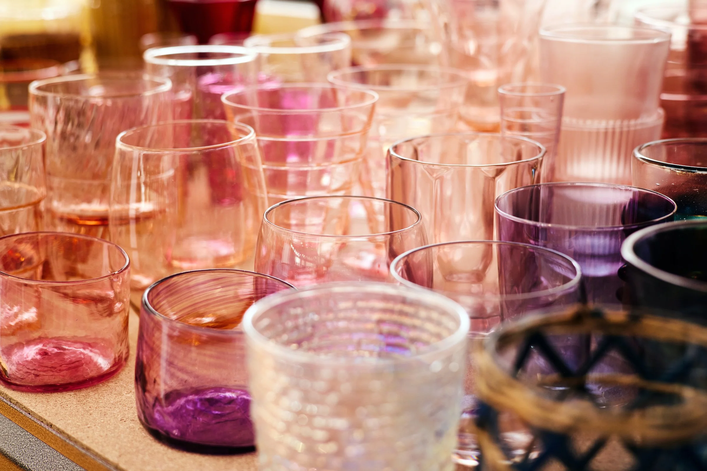 Colorful glassware on a wooden surface, including various sizes and shapes of tinted glass cups in shades of pink, purple, and orange, arranged closely together.
