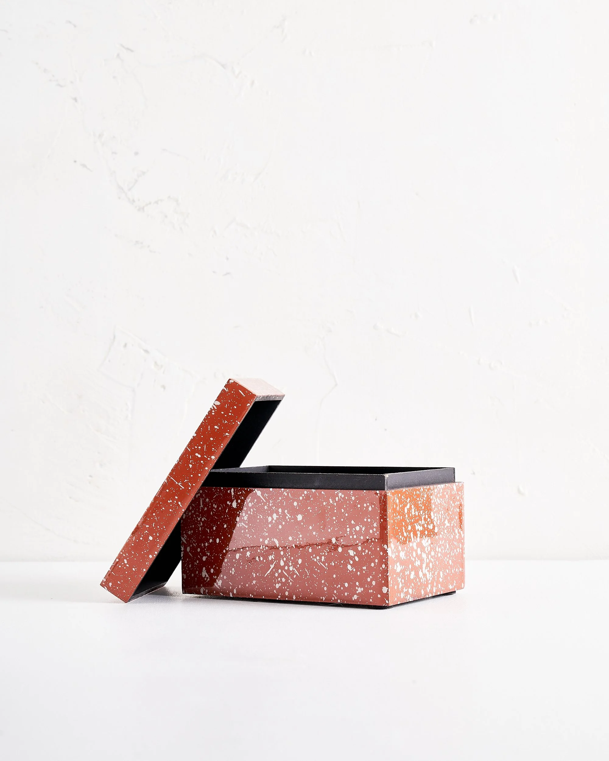 A rectangular terracotta colored speckled box with a lid leaning against it, sitting on a white surface against a white textured wall.