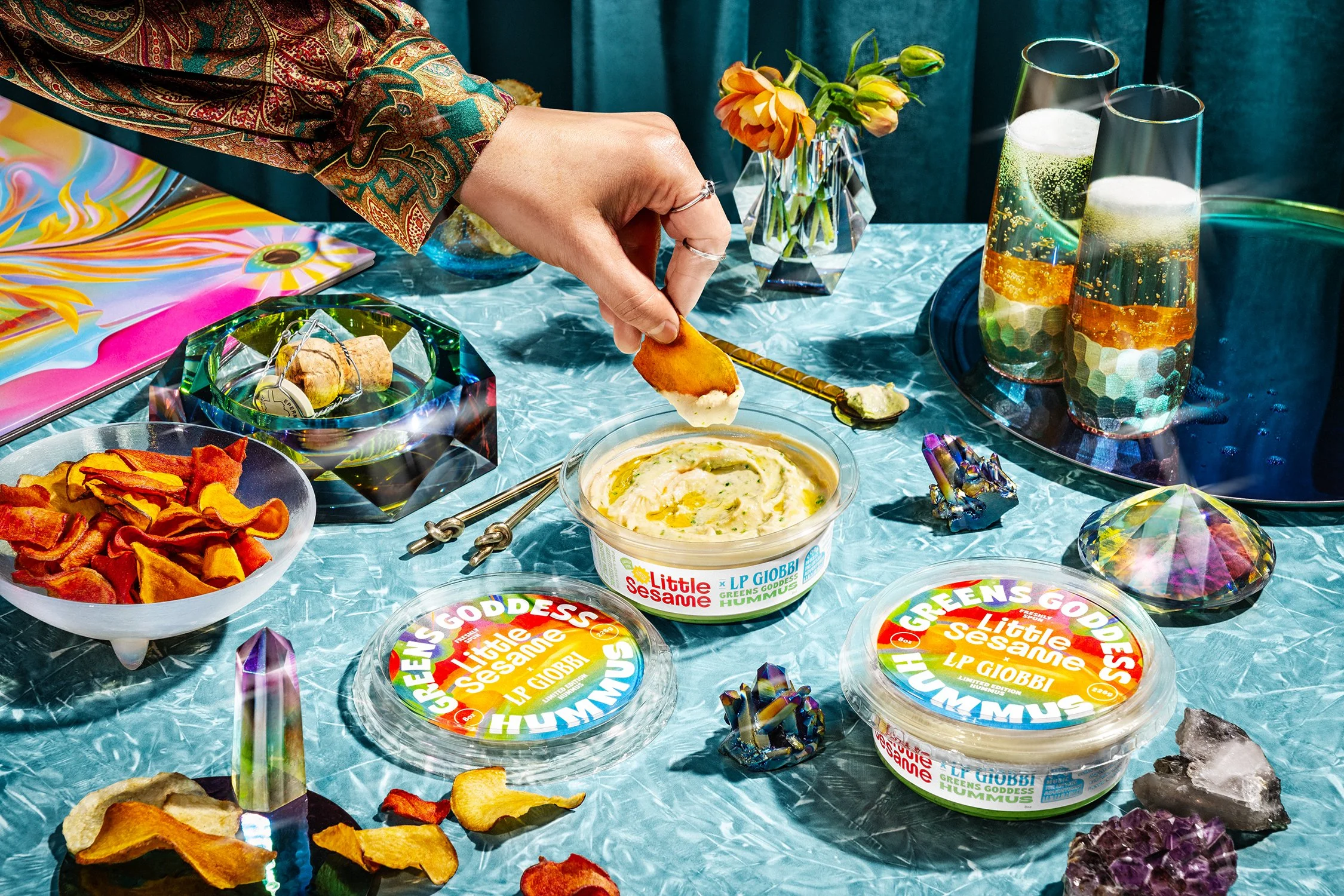 A hand dips a chip into a container of "Green Goddess" hummus on a table with colorful decor, flowers, and glasses with drinks. Nearby are vibrant gemstones and a bowl of chips.