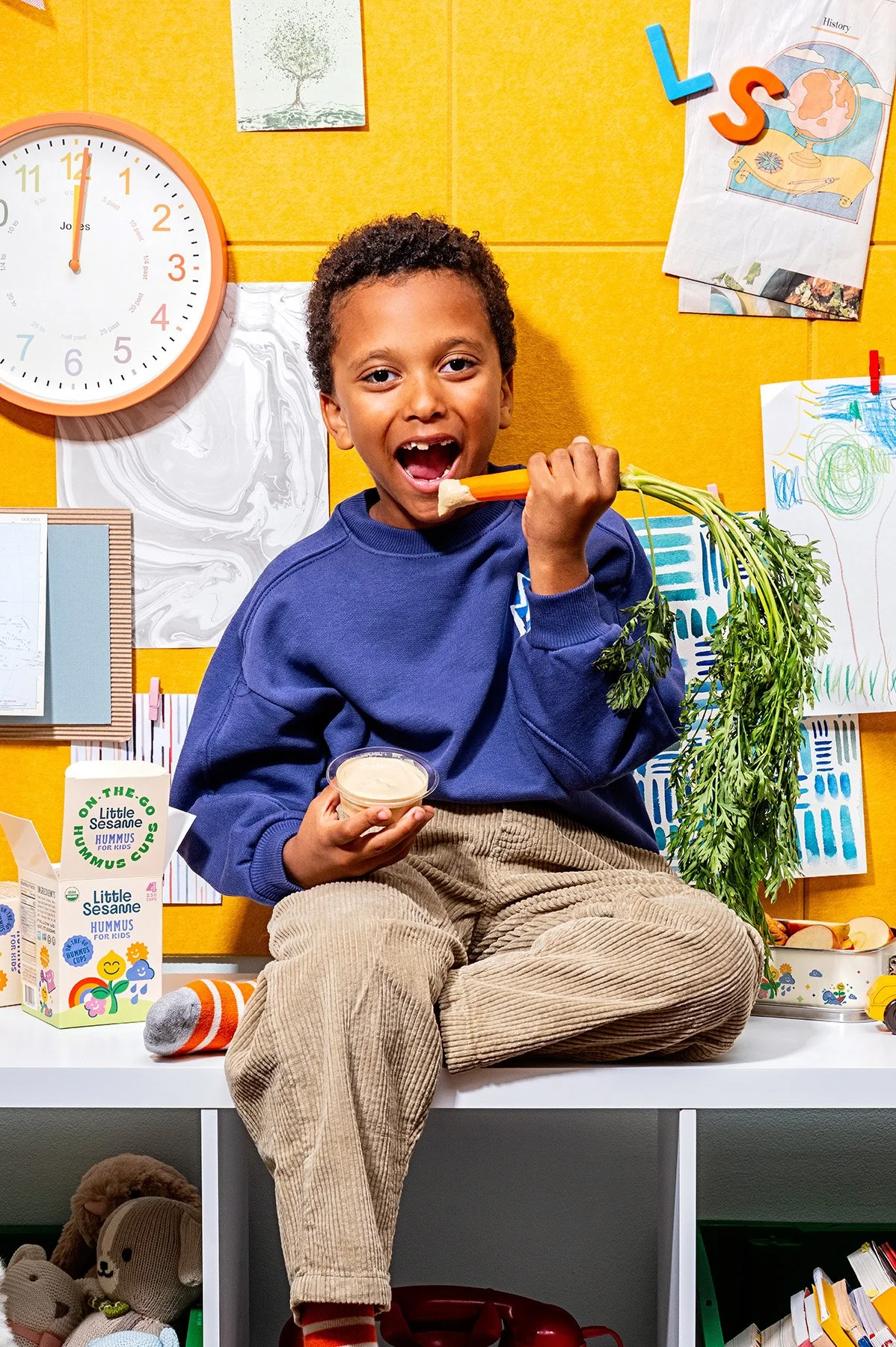 Child eating carrot with hummus, sitting on shelf, surrounded by bright yellow background, wall art, clock, and toys.