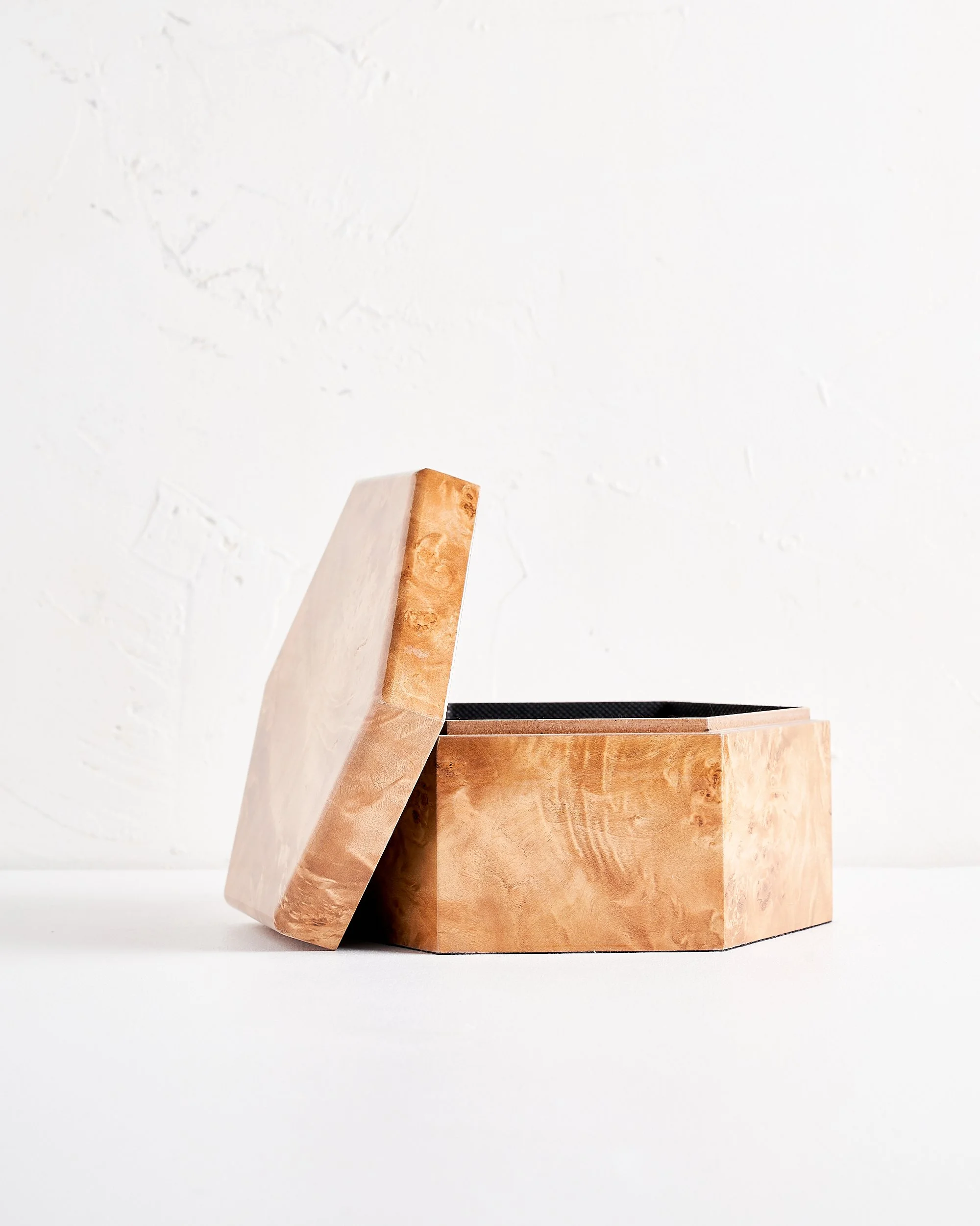 A shallow hexagonal wooden box with a lid off next to it, made of light-colored, polished burl wood, placed on a white surface against a white wall.