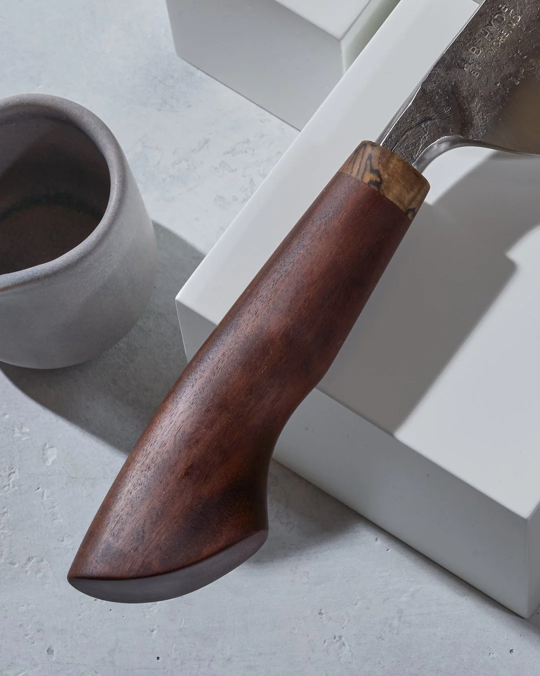 A close-up of a handmade knive with a wooden handle resting on a white surface, next to a gray ceramic cup and a white box.