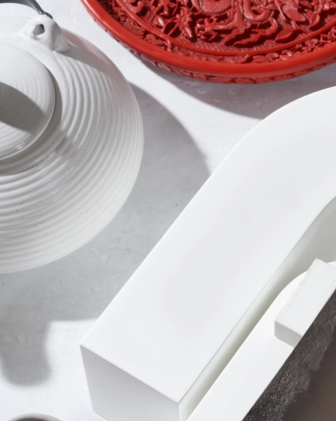 Close-up of a white knife with a black handle, a red round decorative box with intricate cut-out patterns, and a white ceramic teapot with a textured design on a light surface.
