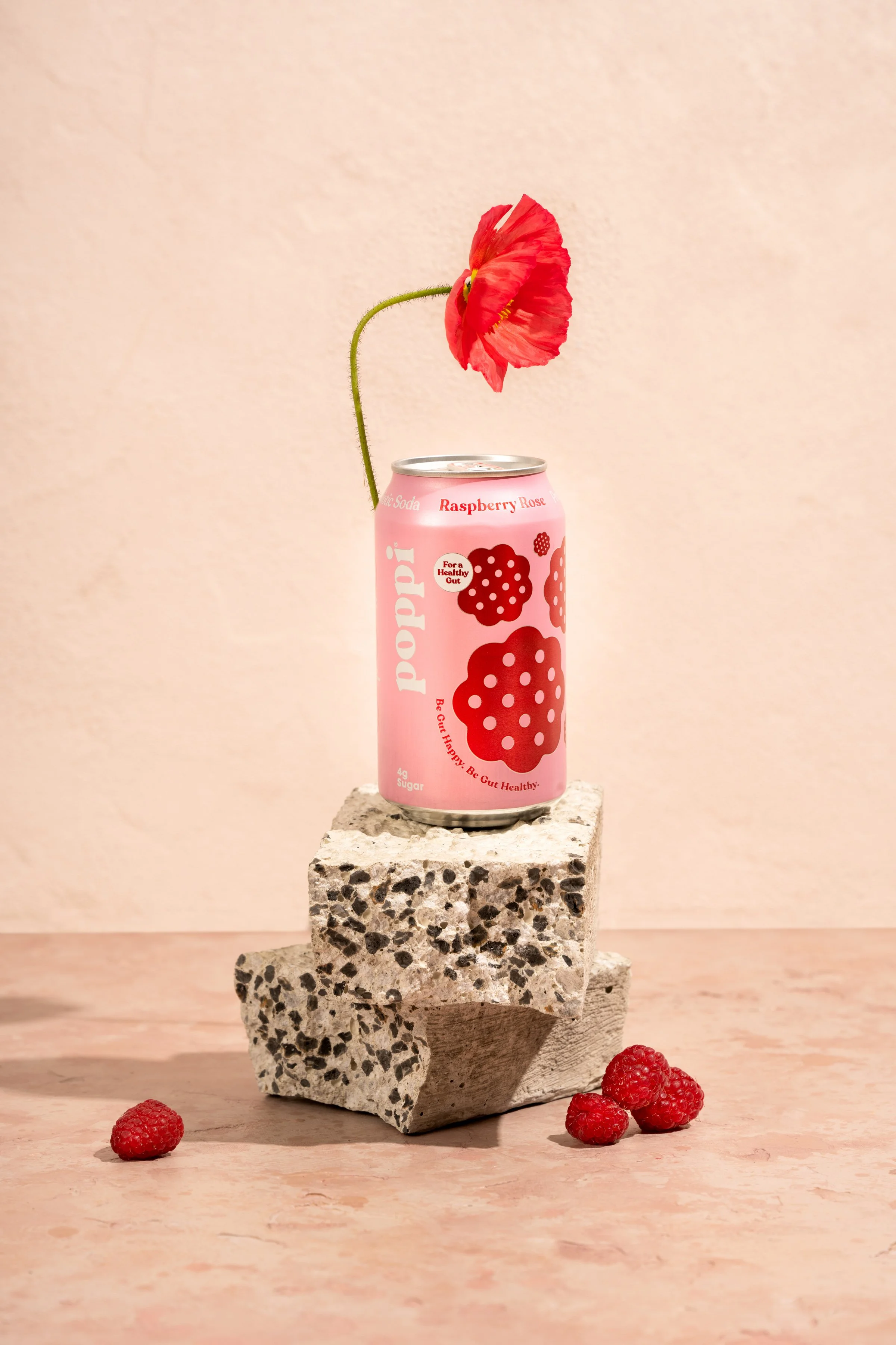 A pink raspberry-flavored soda can labeled 'poppi' with red raspberry illustrations, placed on a concrete block with a red flower and berries around it against a beige wall.