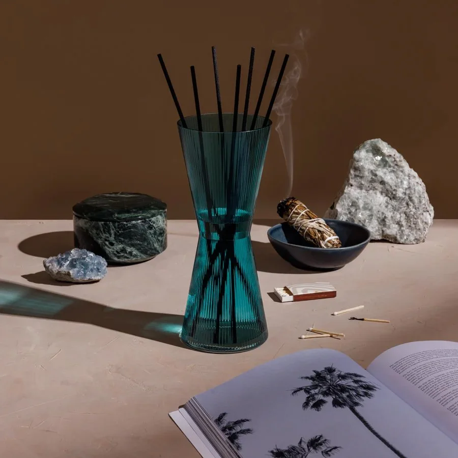 A teal glass vase holds black sticks, surrounded by a green marble container, a piece of blue crystal, and a smoking bundle of sage in a ceramic bowl. Nearby are matches and an open magazine with a palm tree image.