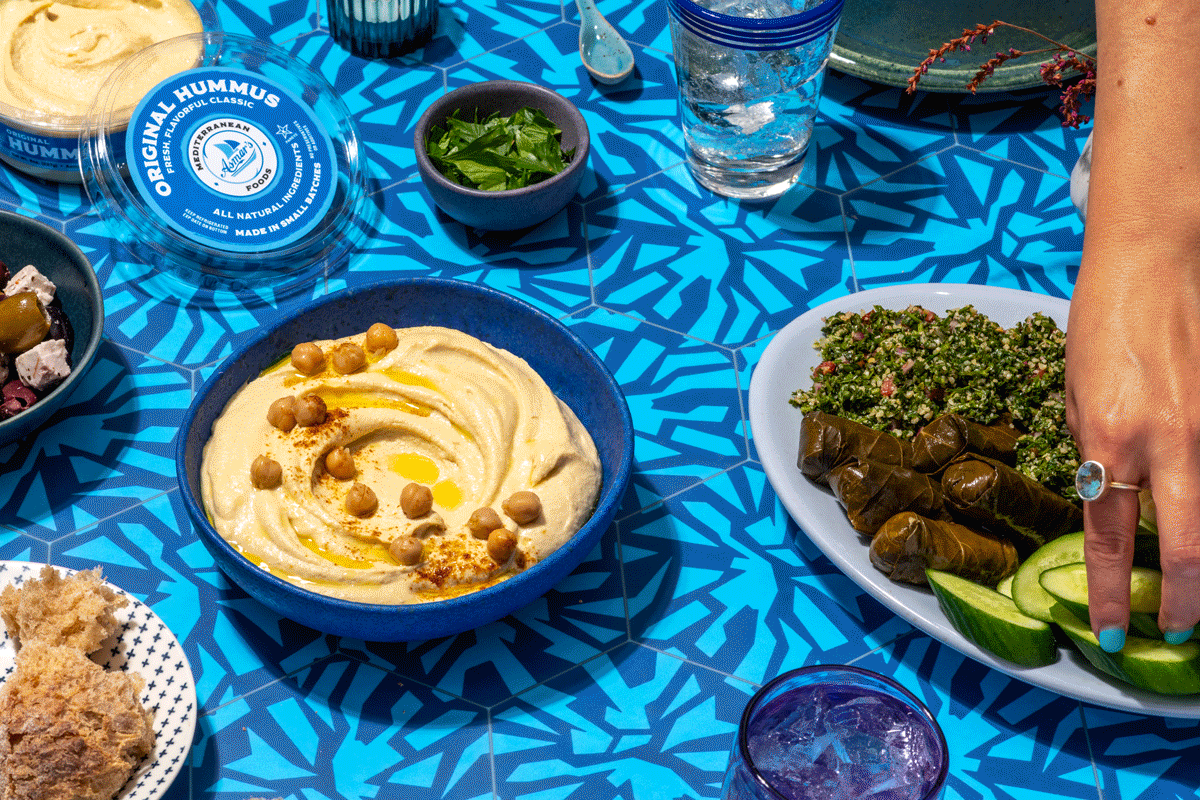 A table with various Middle Eastern dishes, including a bowl of hummus topped with chickpeas and paprika, a plate of stuffed grape leaves and tabbouleh, and a small bowl of fresh herbs, set on a blue patterned tablecloth.