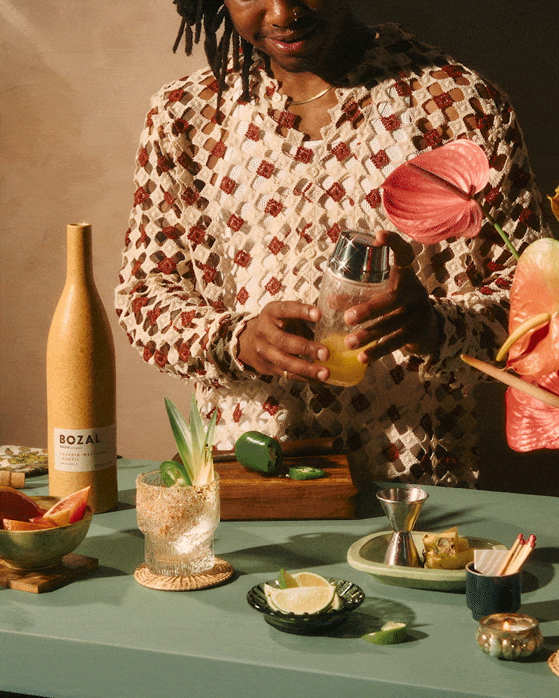 A man preparing mezcal based cocktails with hibiscus flowers and various ingredients on a green table.