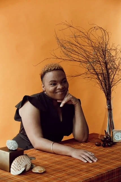 A dark skinned woman with short blonde hair smiling and resting her chin on her hand at a wooden table with decorative objects, a branch in a vase, and a clock against an orange wall.