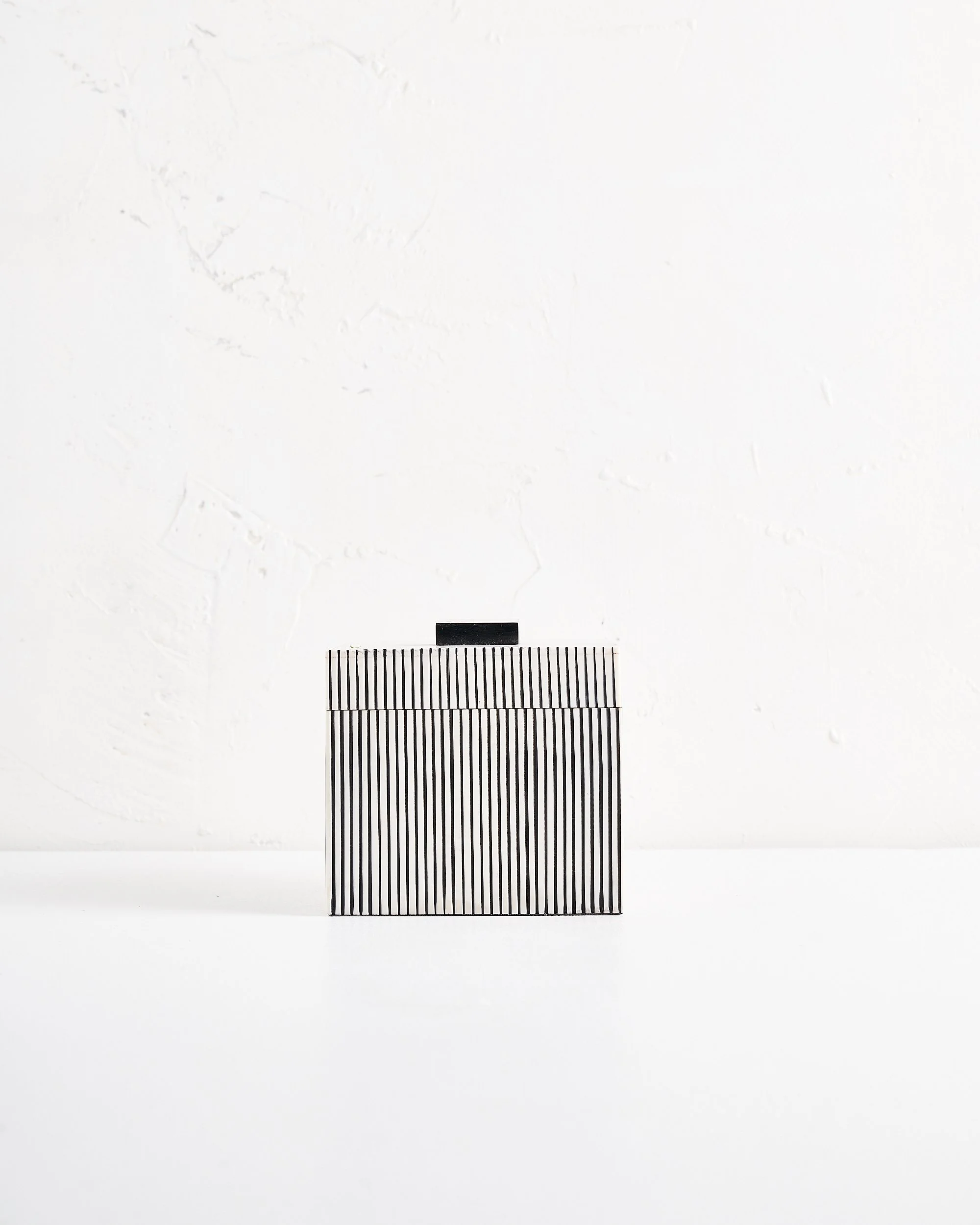 A medium sized modern small square black and white striped box positioned on a white surface against a white wall.