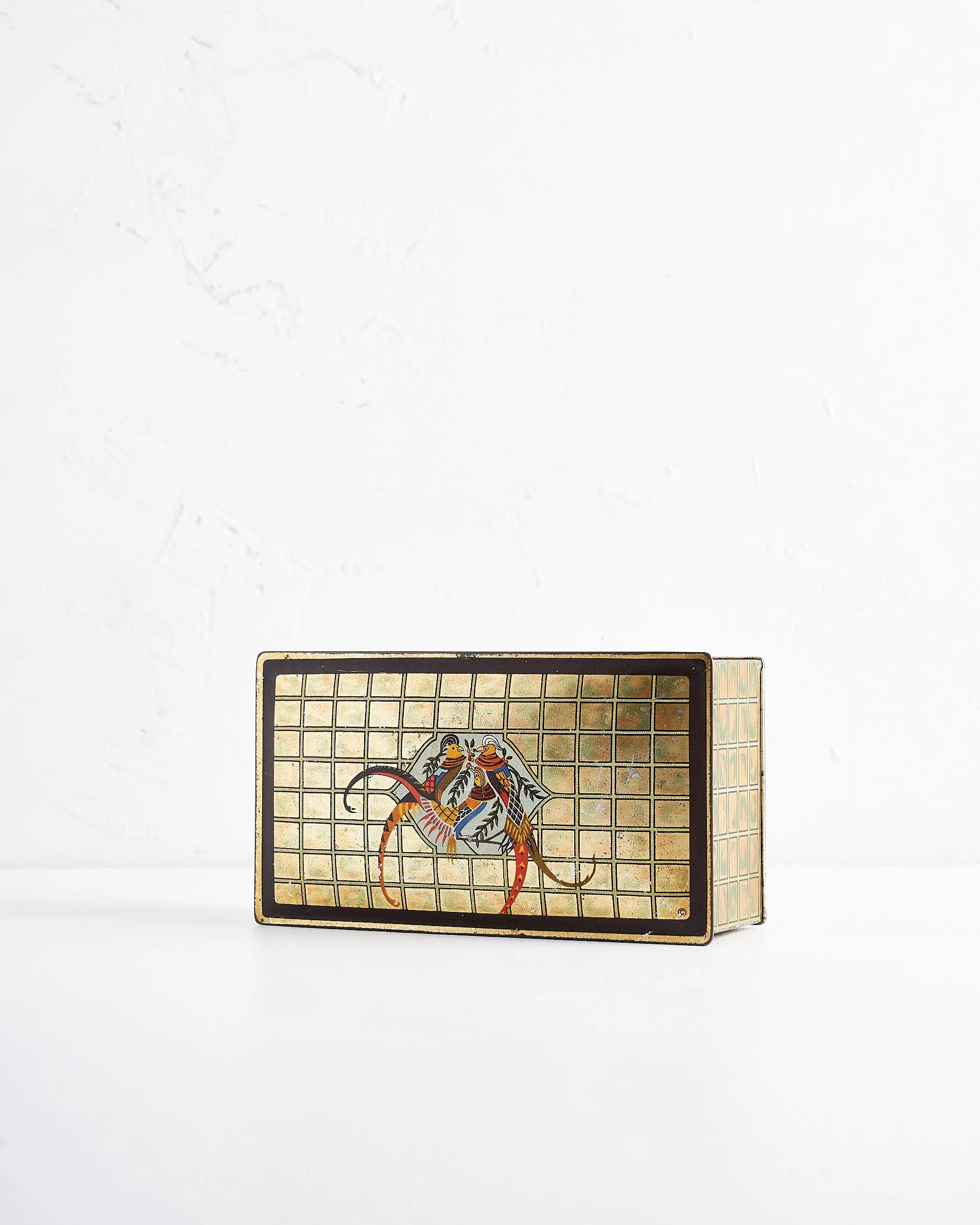 A vintage rectangular metal box with a decorative design of two colorful birds on a gold-colored grid background, placed on a white surface against a white wall.