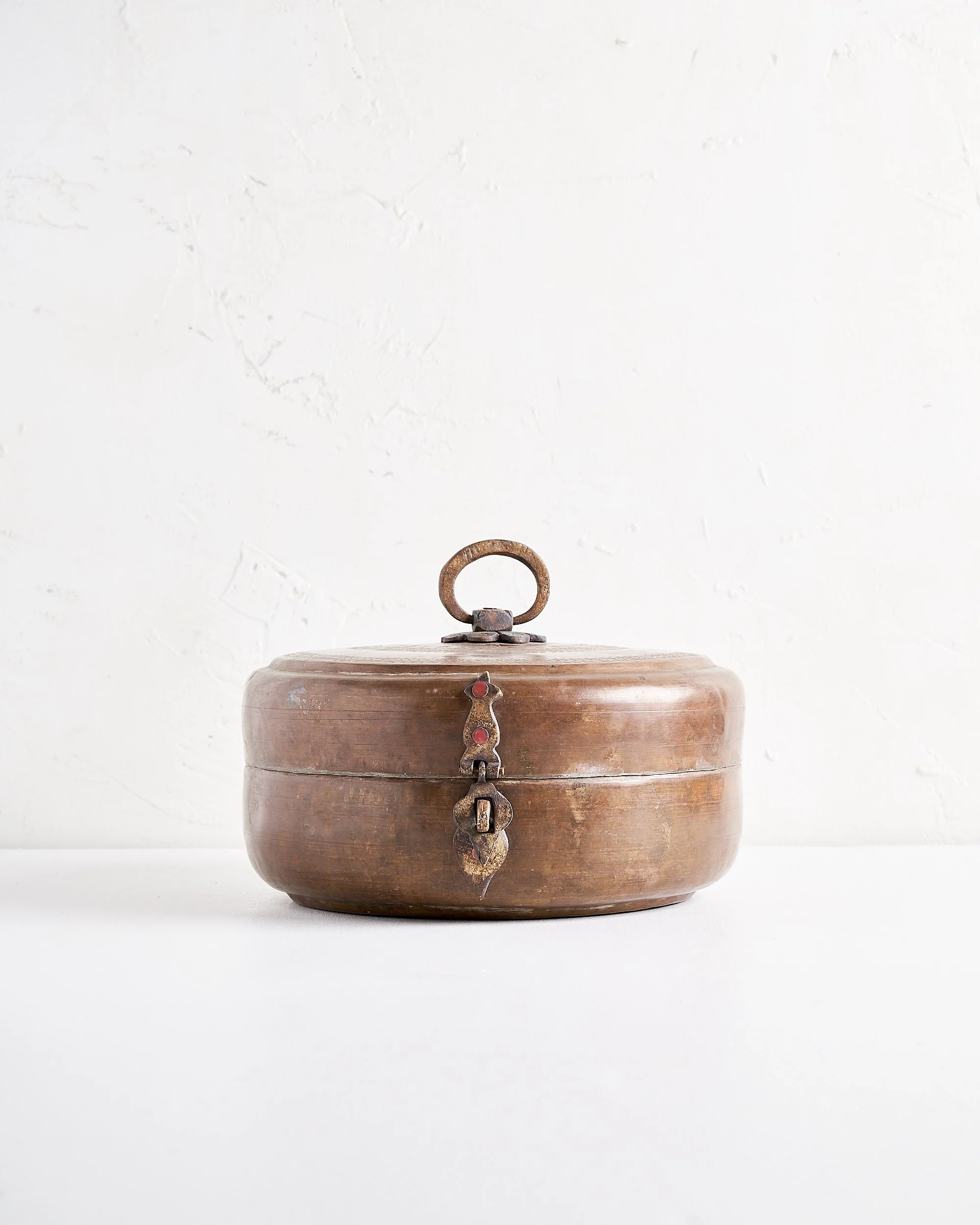 A round, vintage copper metal box with a handle on top and a latch on the front, sitting on a white surface against a white textured wall.