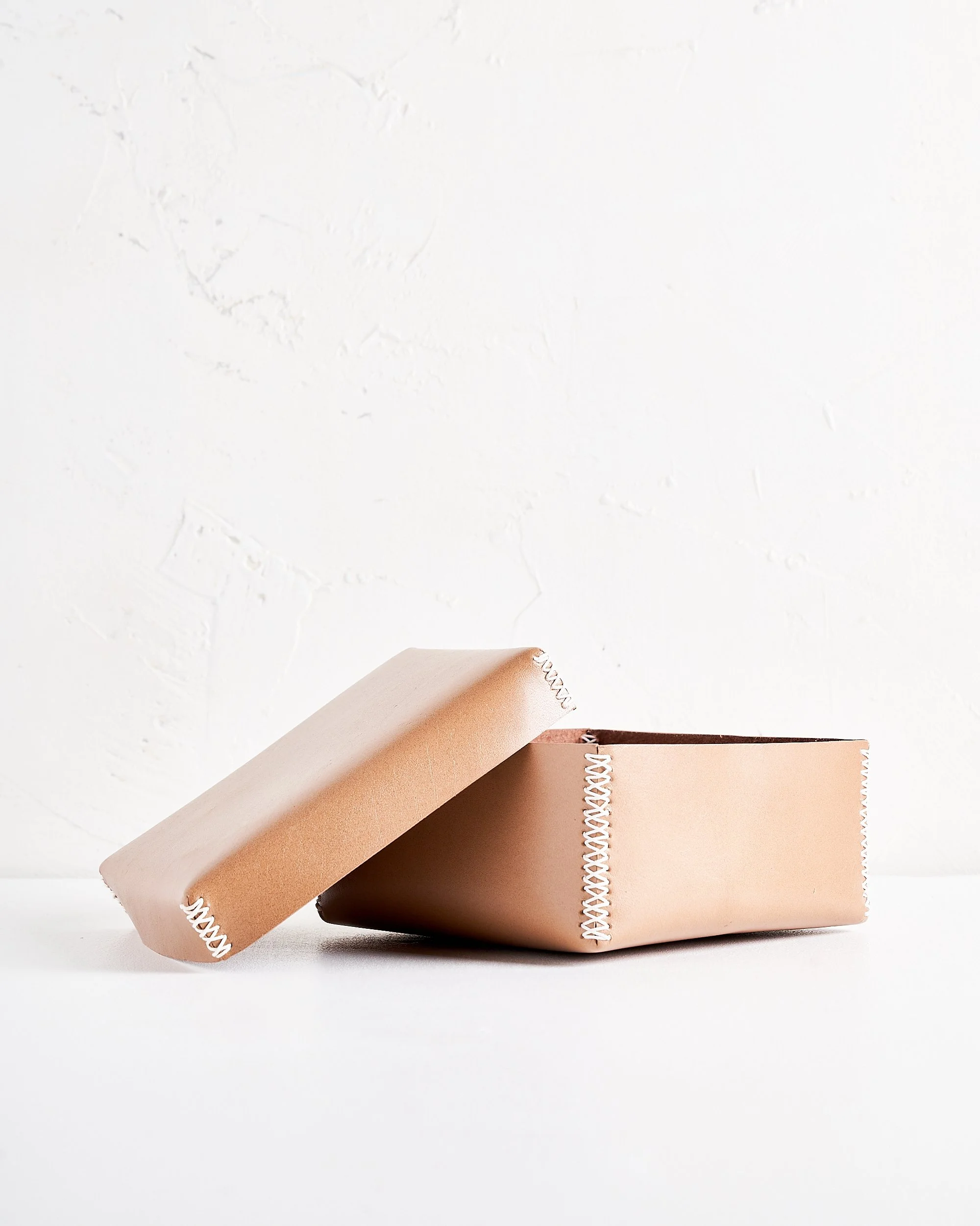 A rectangular tan leather box with white stitched edges with a removable lid, placed on a white surface against a white textured wall, with the lid leaning against the box.