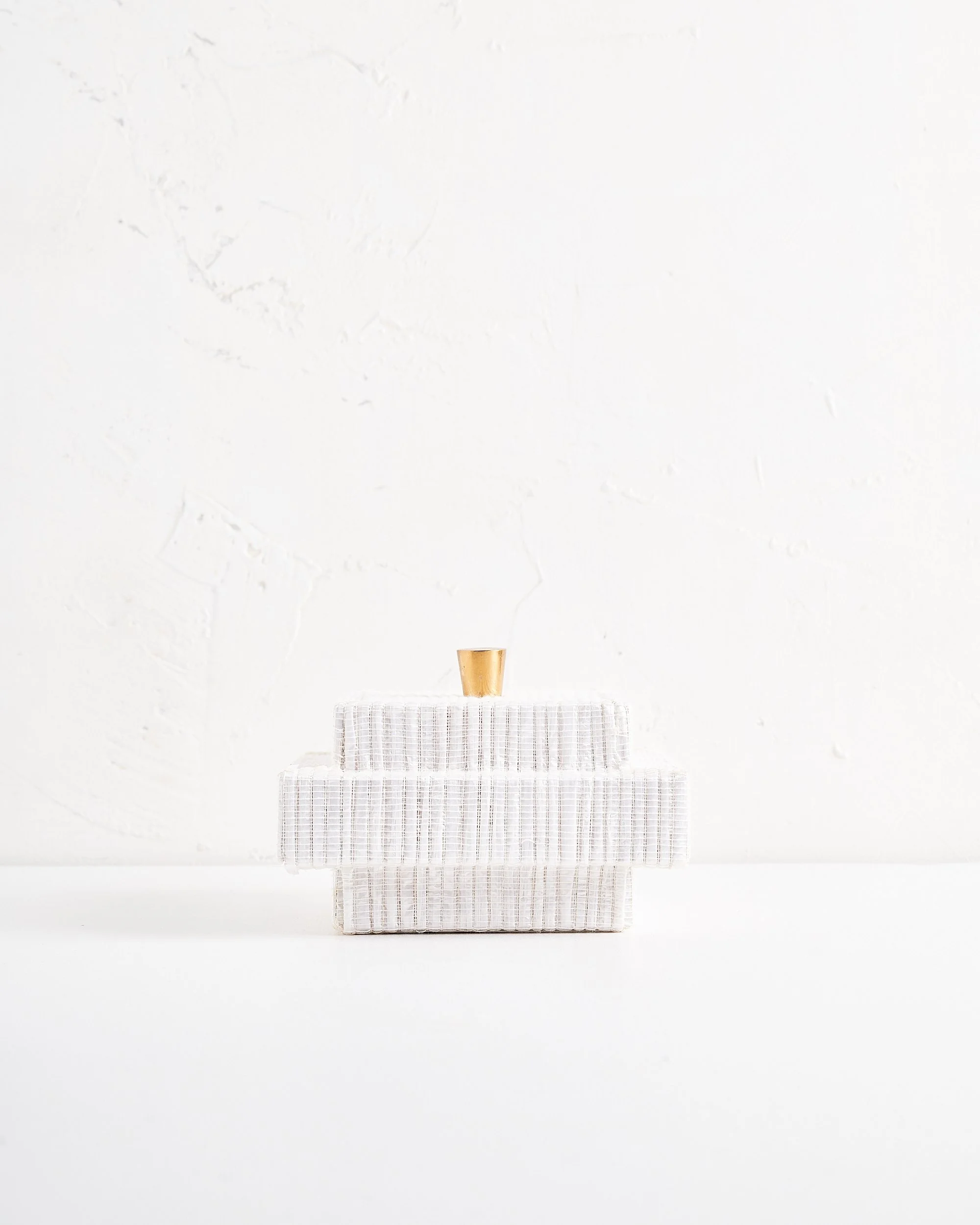 A modern decorative white fabric jewelry box with minimalist brass handle on lid, placed on a white surface and textured white wall.
