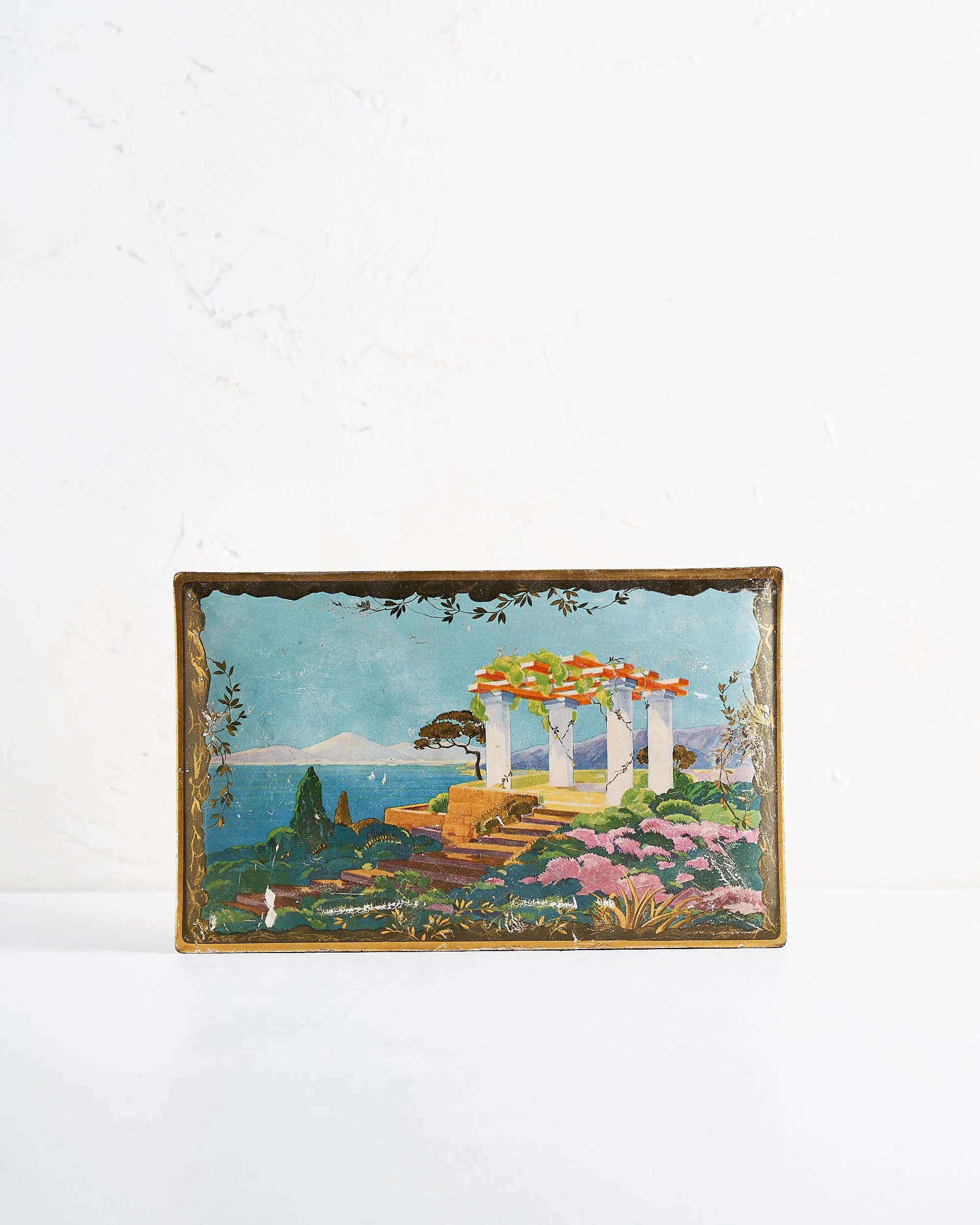 A metal box showing lid with a colorful vintage landscape illustration of a seaside scene with a Greek-style temple, stairs, lush flowers, and mountains in the background. Placed on a white surface against a white textured wall.