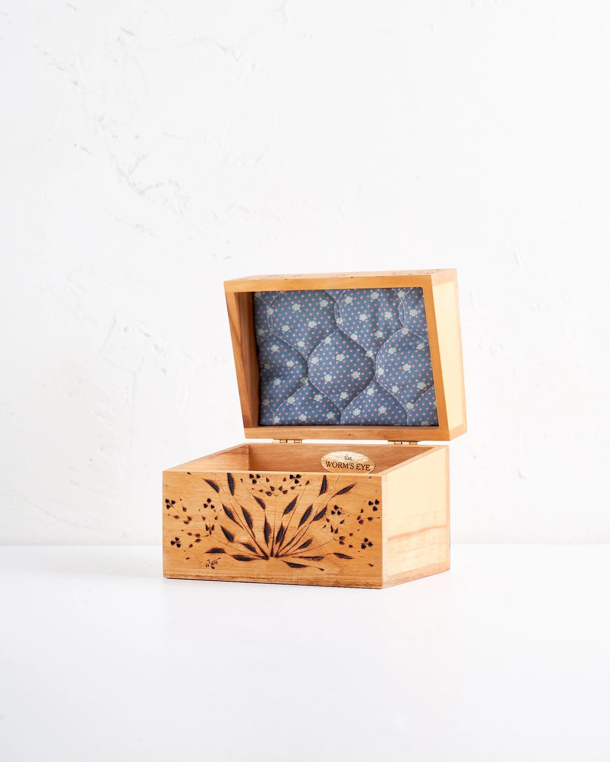 An open tan antique handmade wooden box with a dark stained floral design carved on the front, placed on a white surface against a white textured wall.