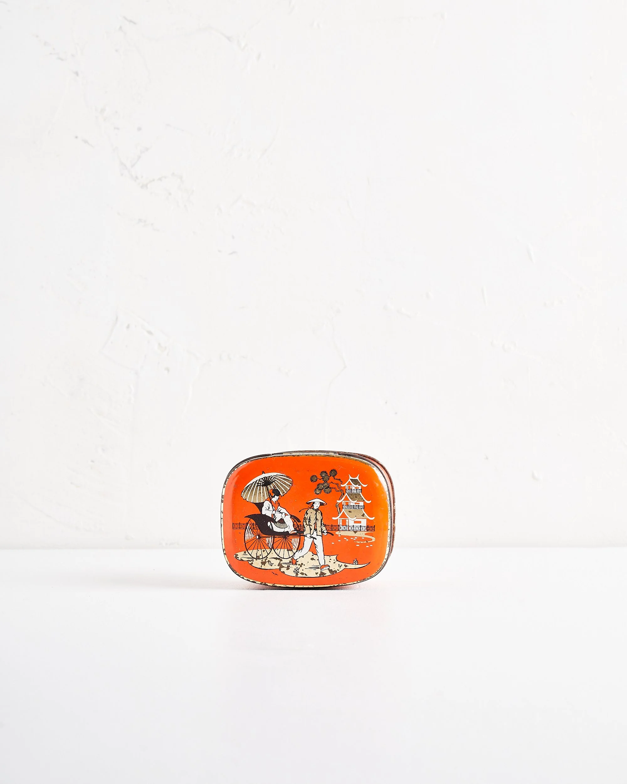 A small rectangular tin with an orange background, featuring a vintage illustration of a man and woman in traditional Asian attire, with a pagoda and landscape in the background.