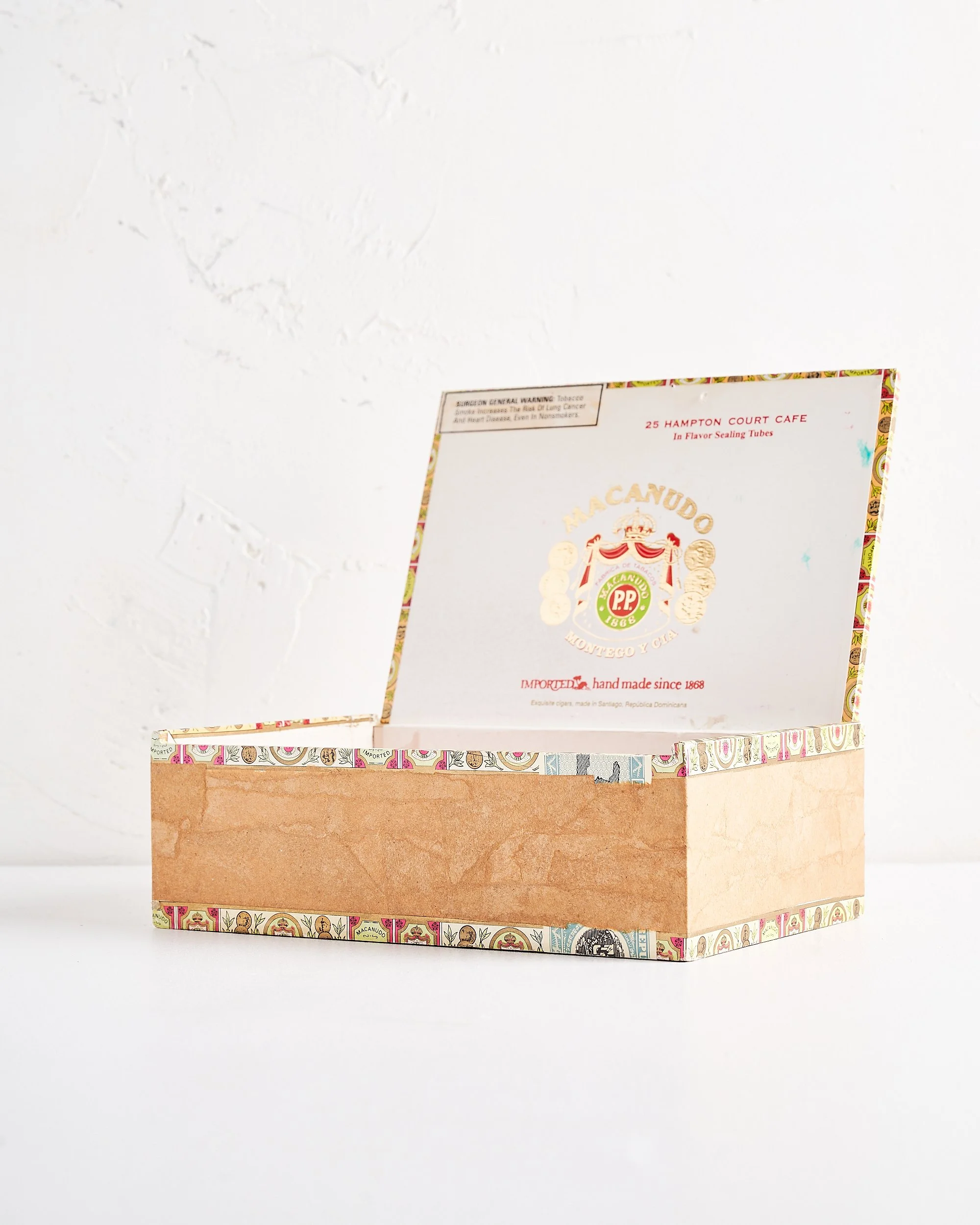 An open rectangular cigar box with a plain brown paper exterior and colorful decorative border on top, placed on a white surface against a textured white wall.