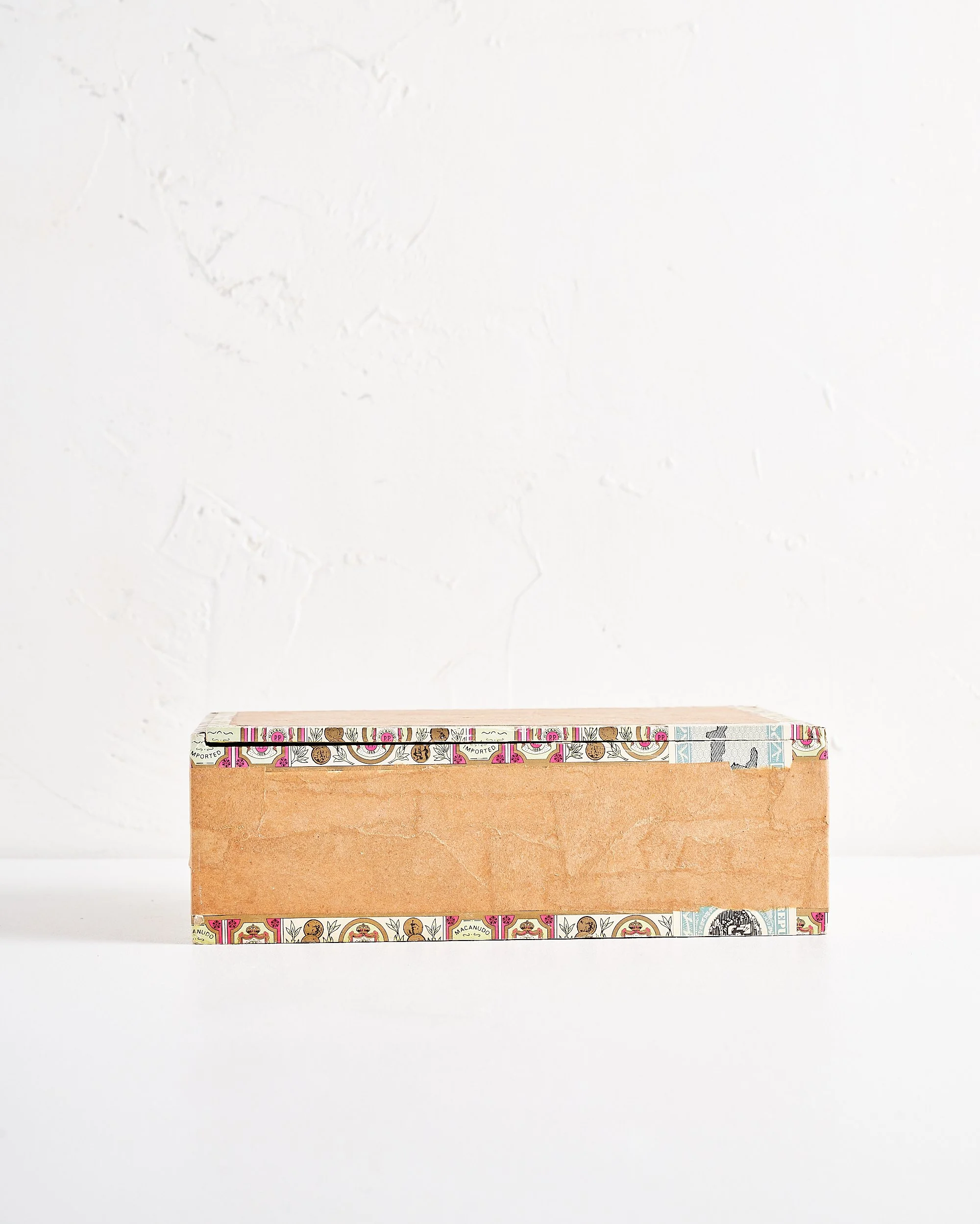A rectangular cigar box with a plain brown paper exterior and colorful decorative border on top, placed on a white surface against a textured white wall.