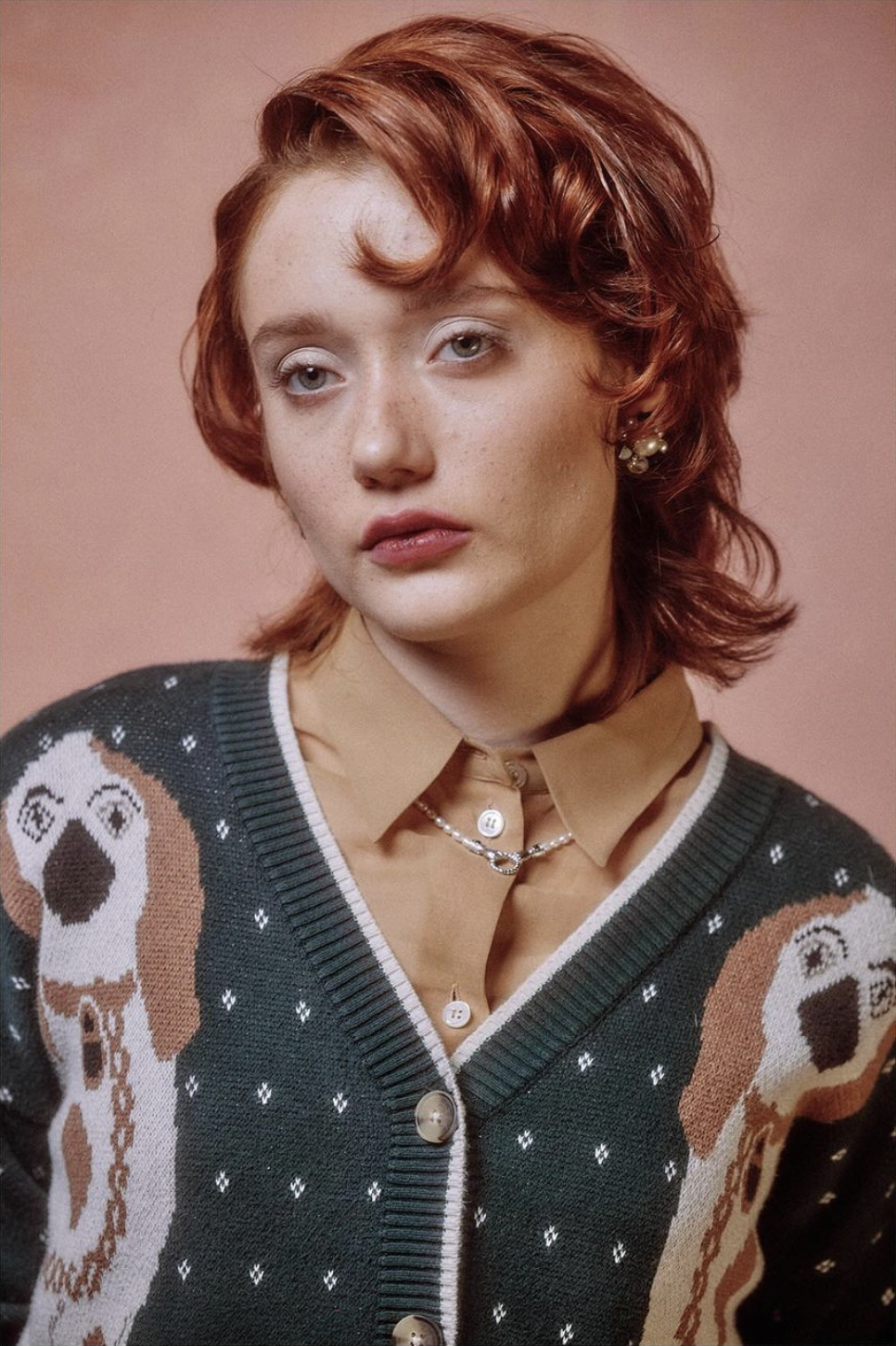 A model with red hair wearing a patterned cardigan and a beige shirt, against a pink background.
