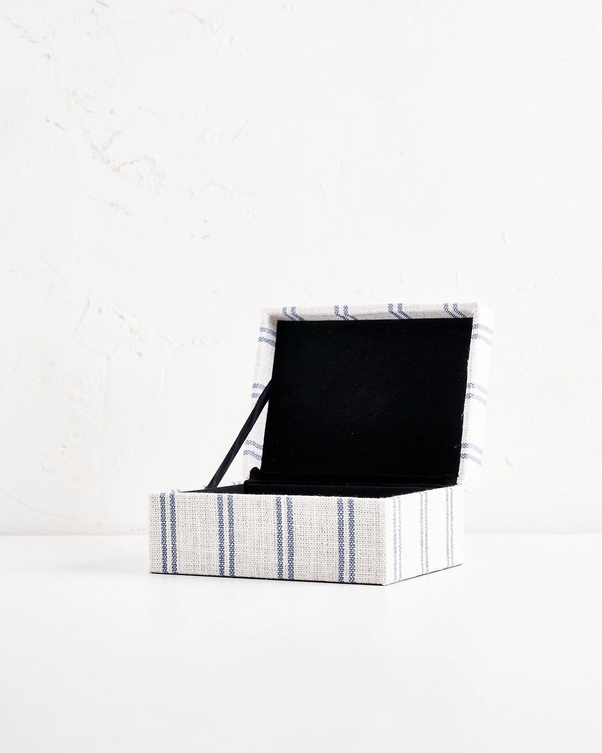 An empty jewelry box with a black interior and a fabric exterior with blue and white stripes, open on a plain white surface against a white wall.