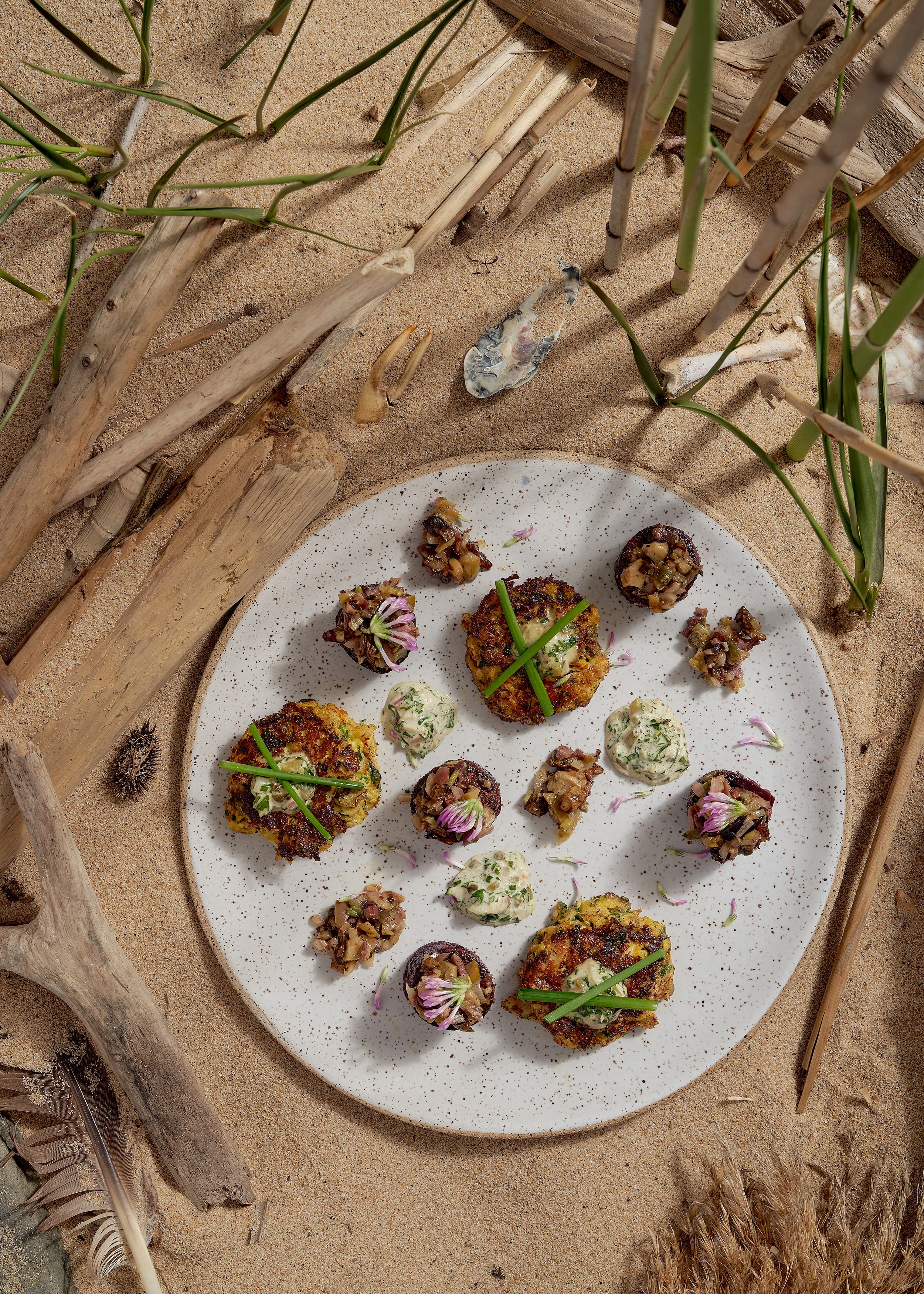 Gourmet appetizers on a white speckled plate on sand, with driftwood and grass, featuring decorative flowers and herbs.