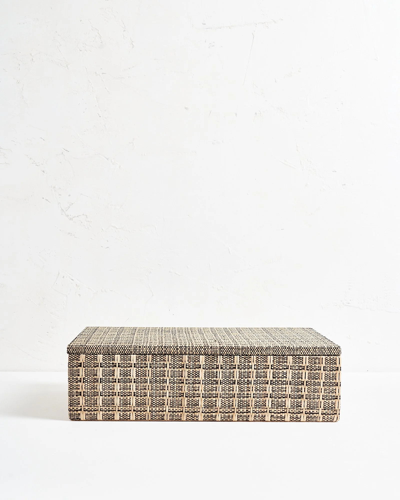 A large decorative rectangular woven black and beige patterned storage box with a cushioned lid, placed against a plain white textured wall.