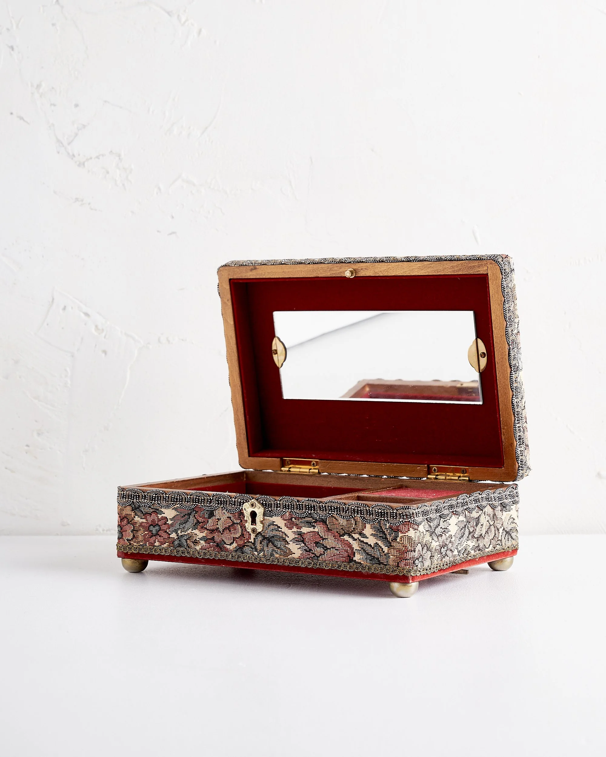Decorative jewelry box with a floral tapestry exterior and a mirror inside, open on a white surface against a white wall.