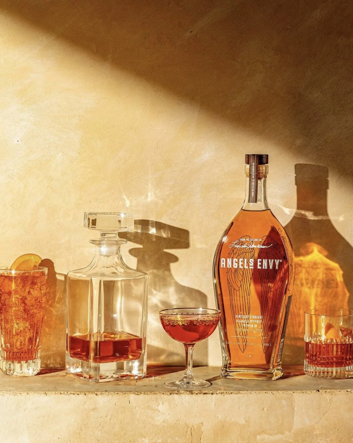 Various bottles and glasses of whiskey on a shelf with sunlight casting shadows on the wall behind them.