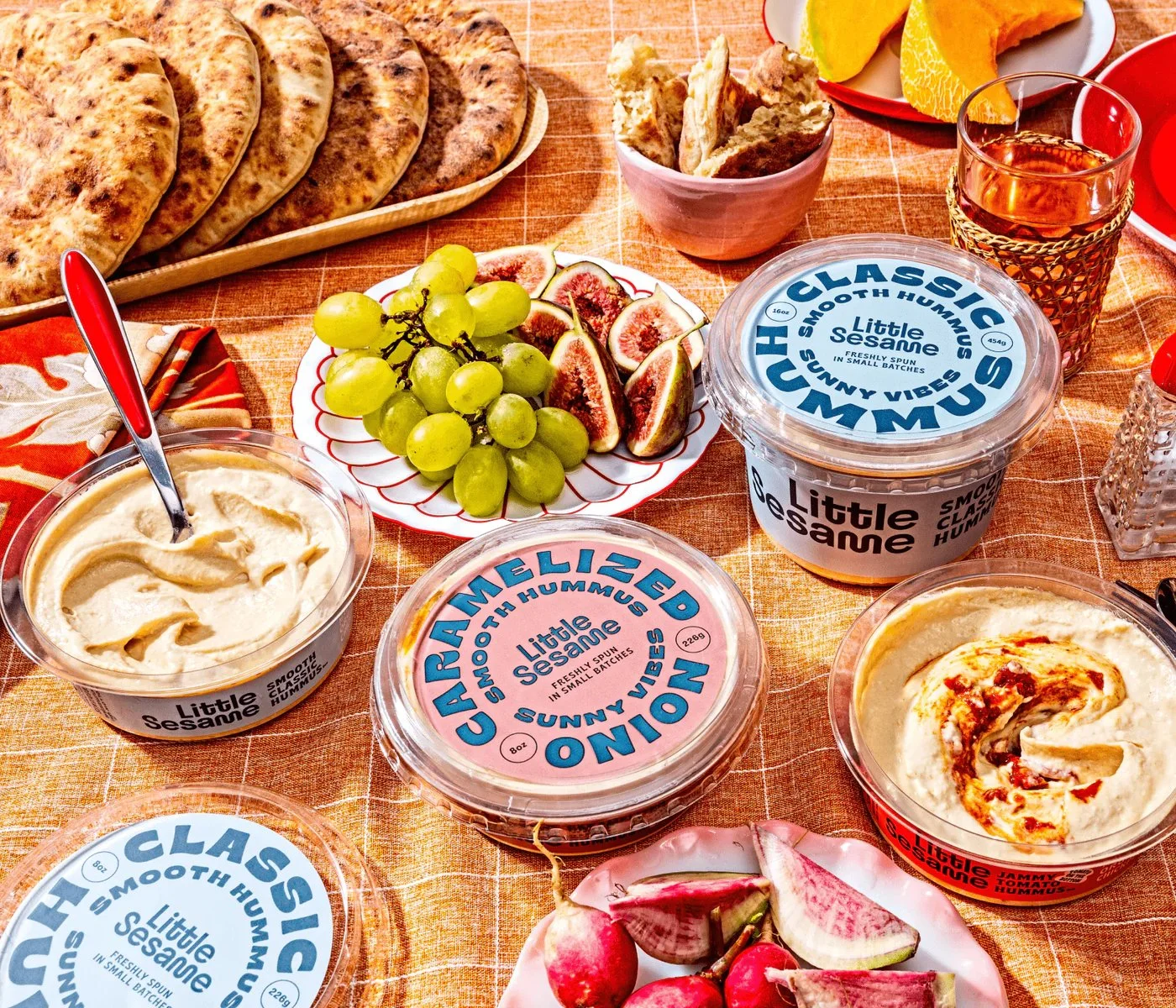 A variety of hummus flavors by Little Sesame displayed on a table with flatbreads, a bowl of figs, green grapes, sliced watermelon radishes, and a glass of drink. The hummus containers have labels such as 'Classic' and 'Caramelized Onion.'
