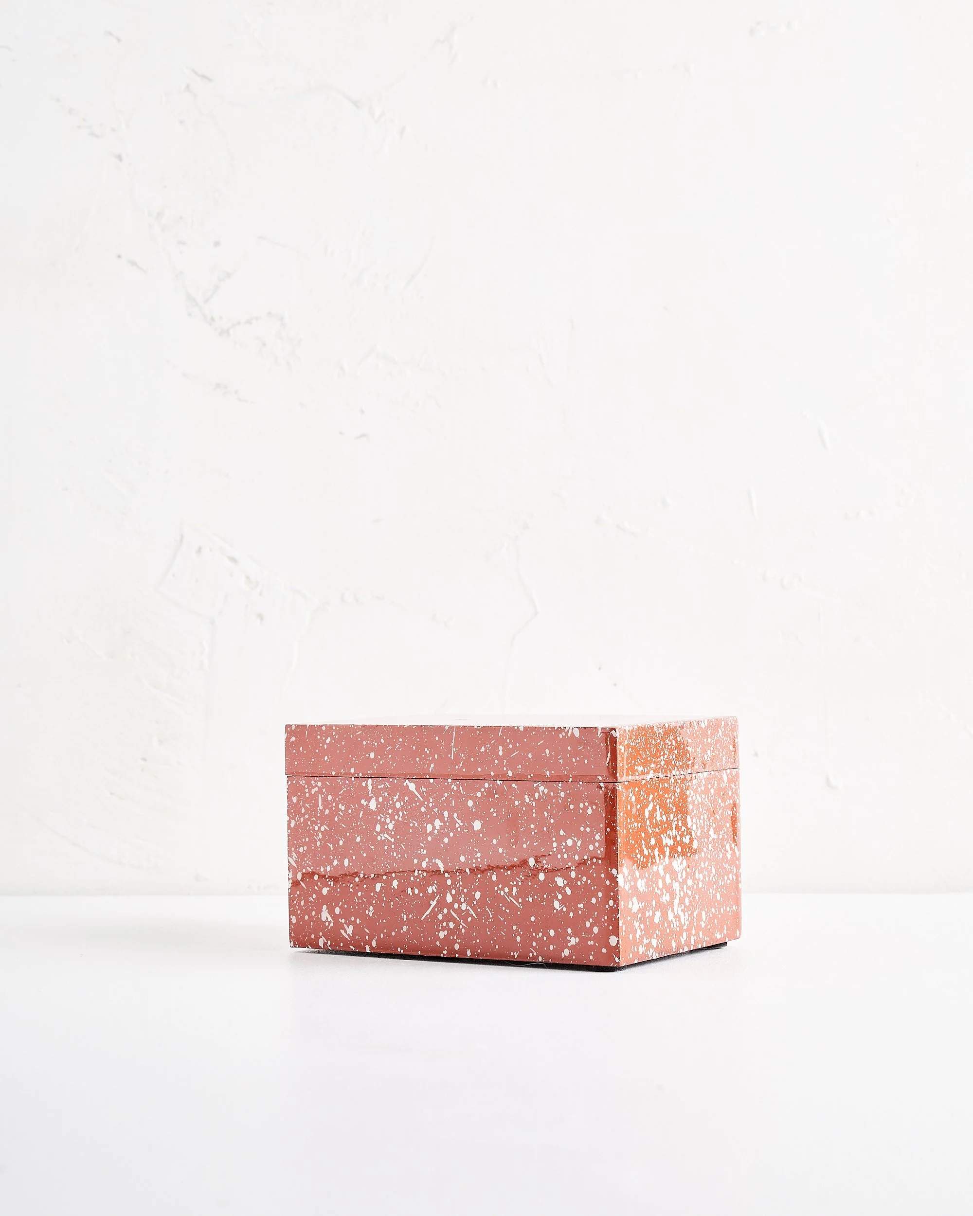 A rectangular terracotta colored speckled box with a lid, sitting on a white surface against a white textured wall.