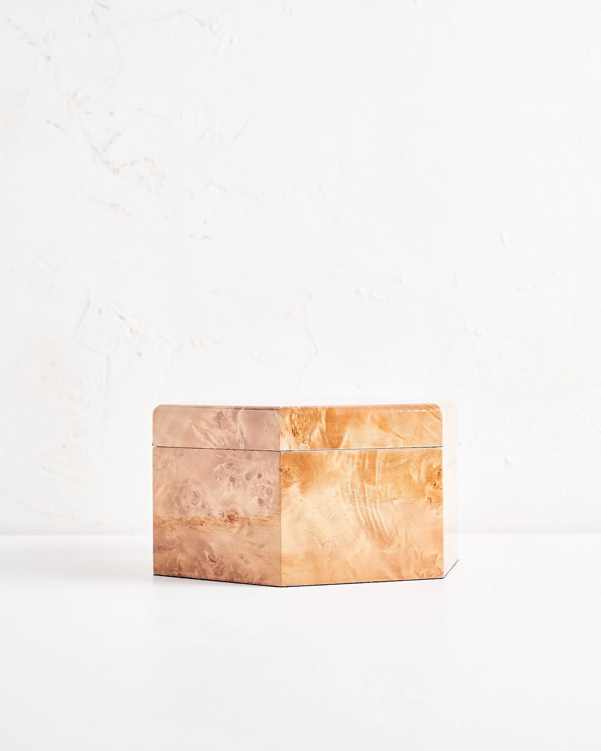 A shallow hexagonal wooden box with a lid, made of light-colored, polished burl wood, placed on a white surface against a white wall.