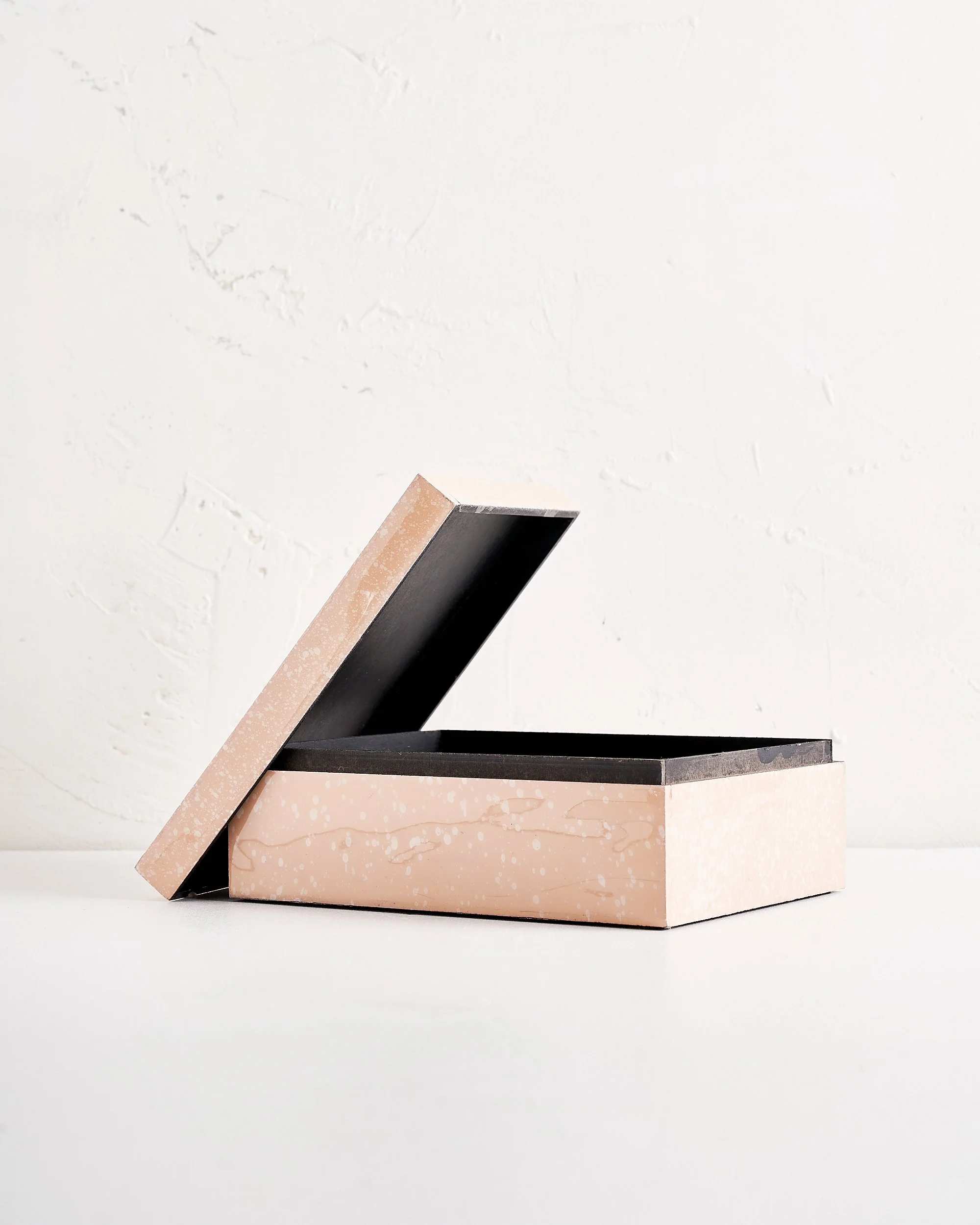 Open rectangular box with a pink exterior and black interior with the lid partly open, placed on a white surface against a white textured wall.
