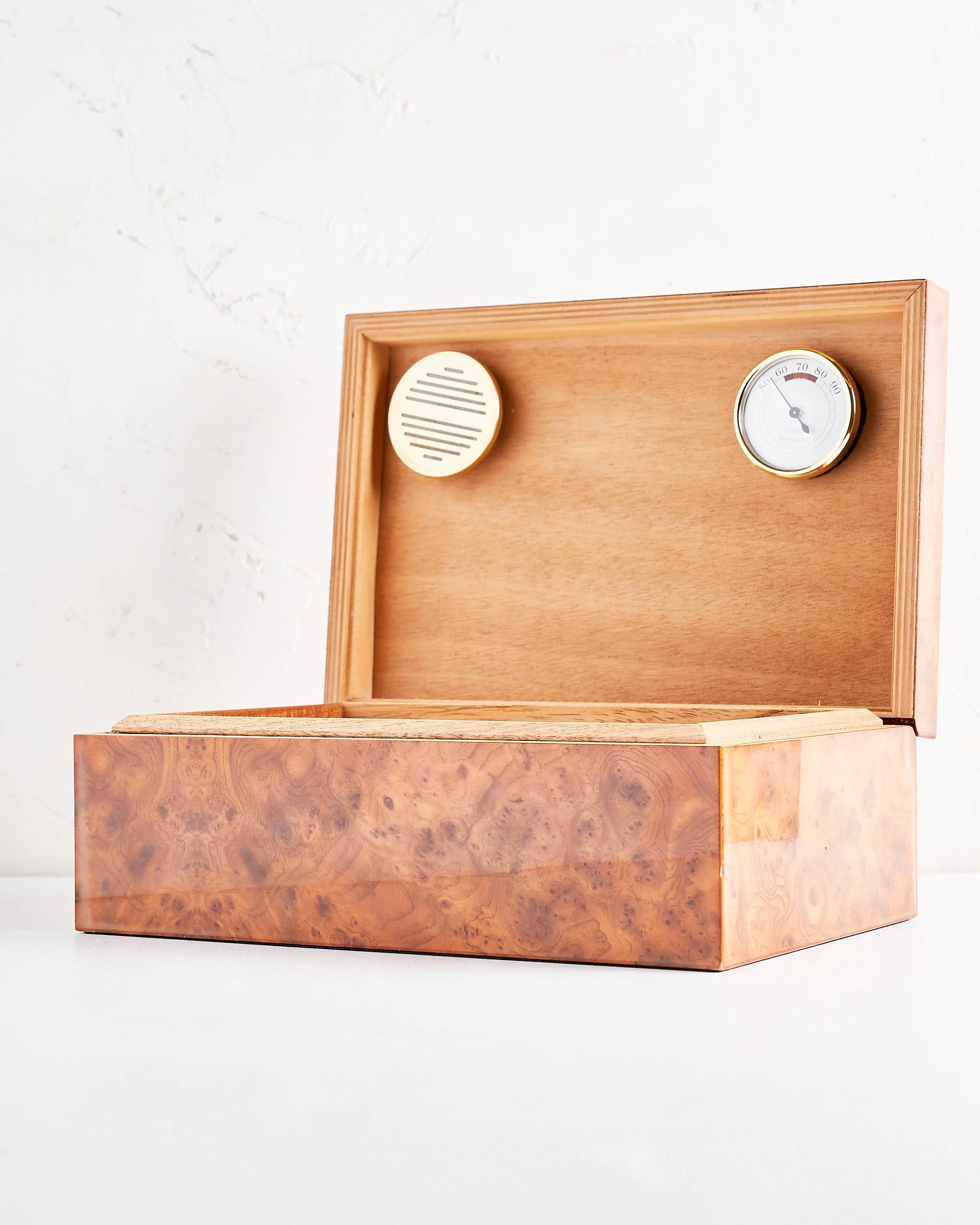 Open wooden cigar box with a ventilation cover and a thermometer inside the lid. Has a light brown, burl wood pattern, placed on a white surface against a white textured wall.