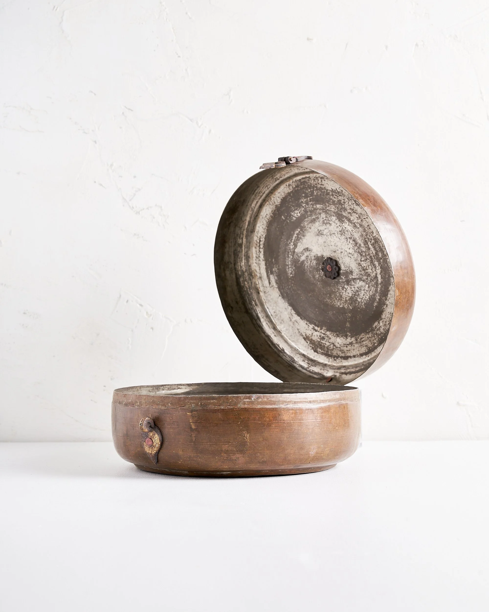 An open, vintage copper and brass tiffin box with a latch, featuring a decorative latch, set against a white textured background.