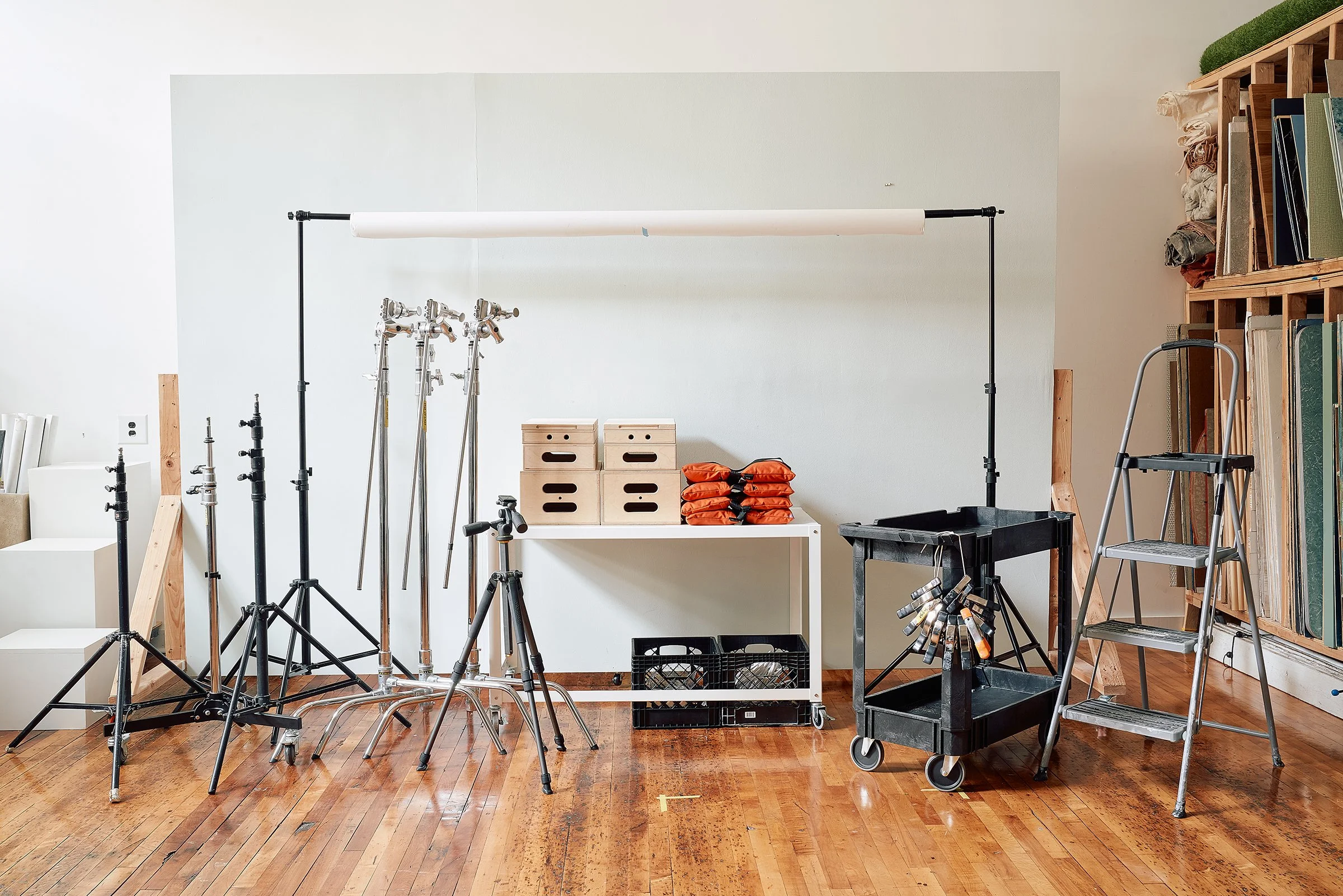 Photography studio equipment including light stands, tripods, C-stands, a rolling cart with sandbags, a stepladder, and a backdrop stand in front of a rolling wall.