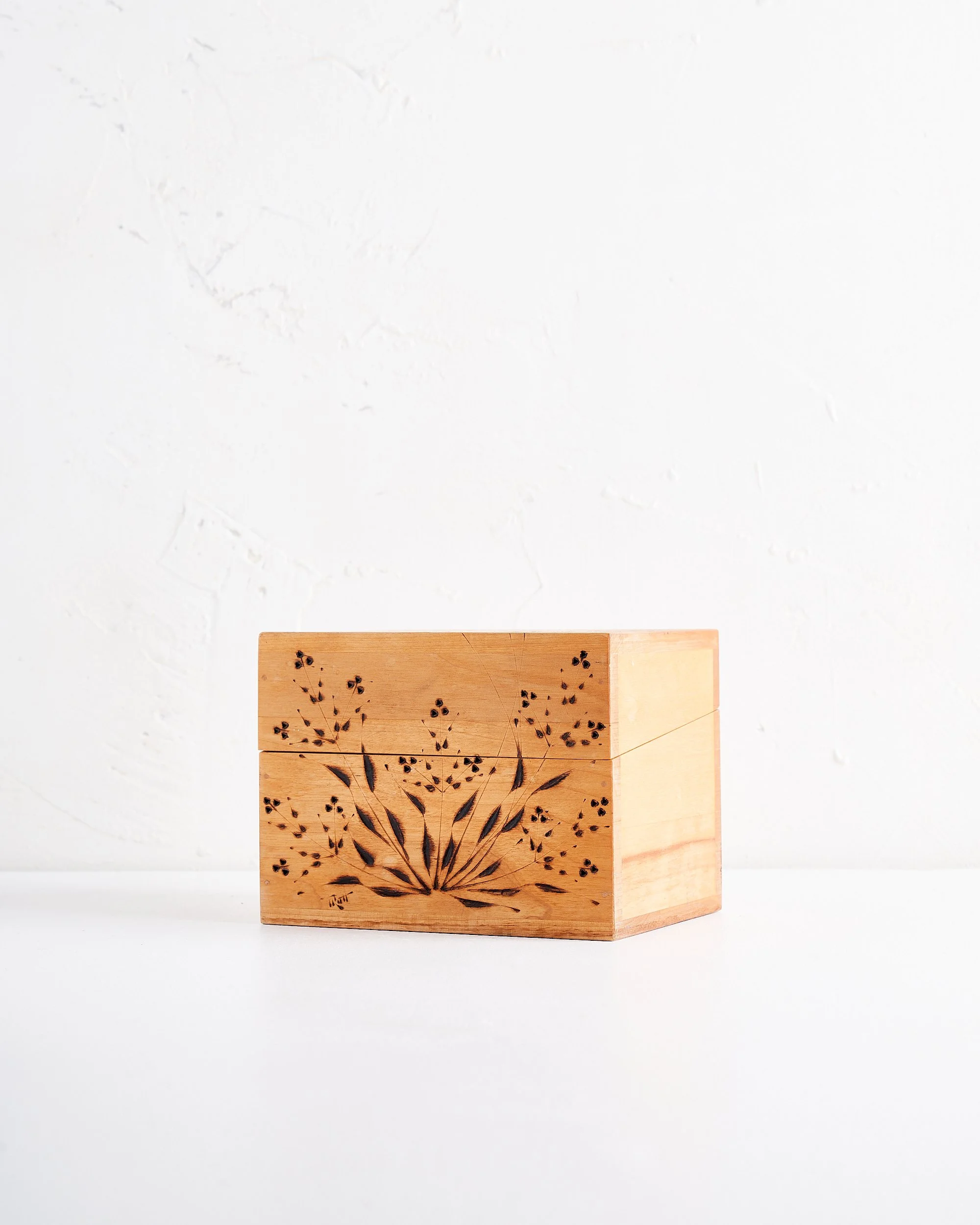 A closed tan antique handmade wooden box with a dark stained floral design carved on the front, placed on a white surface against a white textured wall.