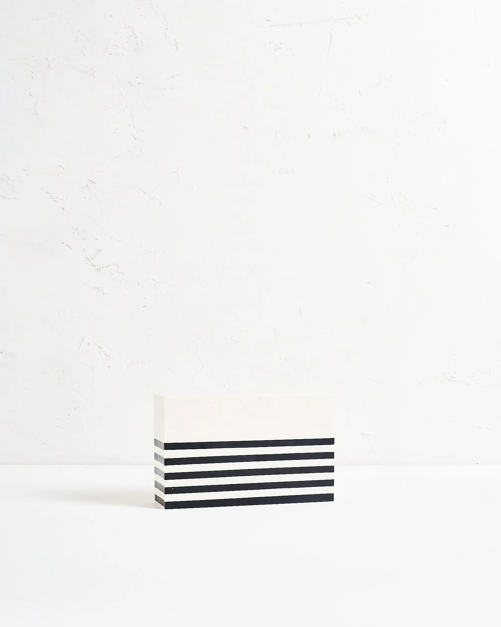 A modern white rectangular box lid with five black horizontal stripes on the lower half, placed on a white surface against a white textured wall.