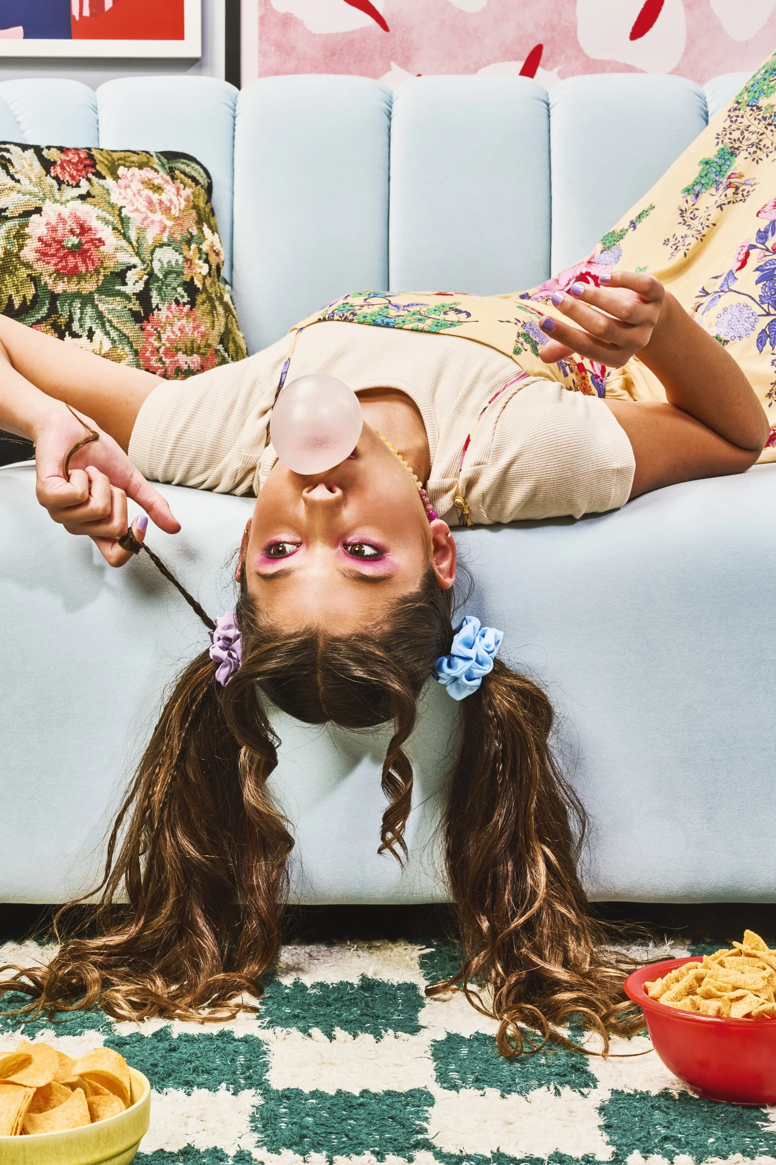 A girl with pigtails lying upside down on a light blue sofa, blowing a bubble gum bubble, with snack bowls on the floor nearby.