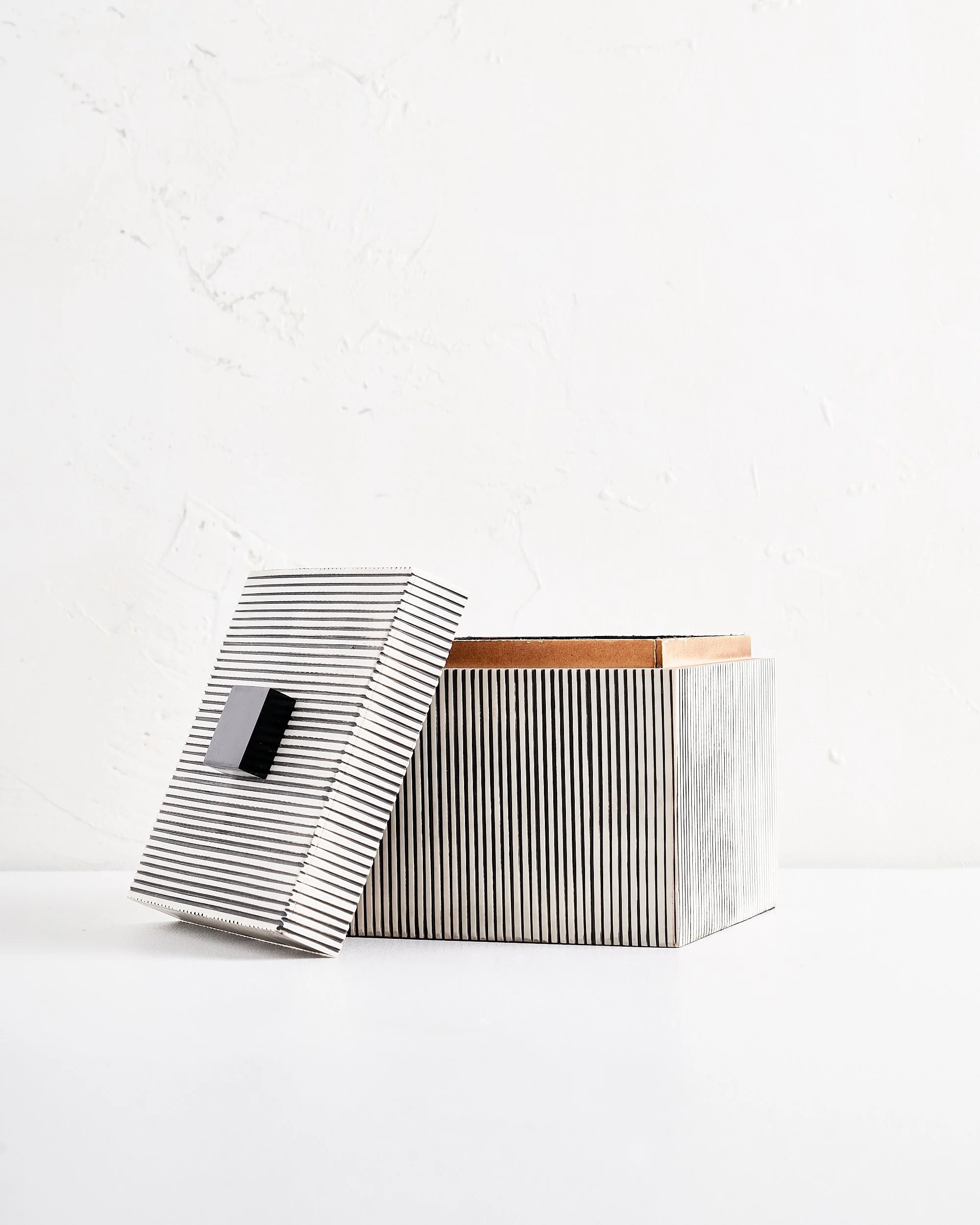 A large sized modern box with black and white vertical stripes on the sides and a lid with matching black and white stripes leaning against it, placed on a white surface agains a white textured wall.