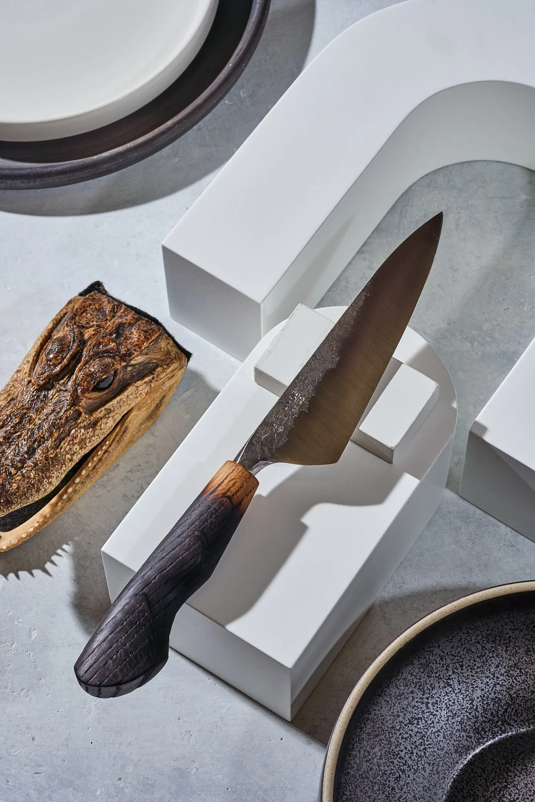 A handmade kitchen knife with a dark wooden handle resting on a white display stand, with a taxidermy alligator head next to it and white and dark bowls in the background.