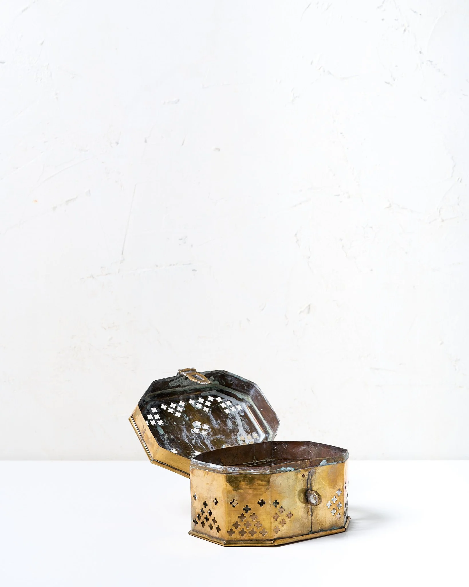 An antique small brass box with a handle, latch, perforated sides and open lid, sitting on a white surface against a plain white wall.