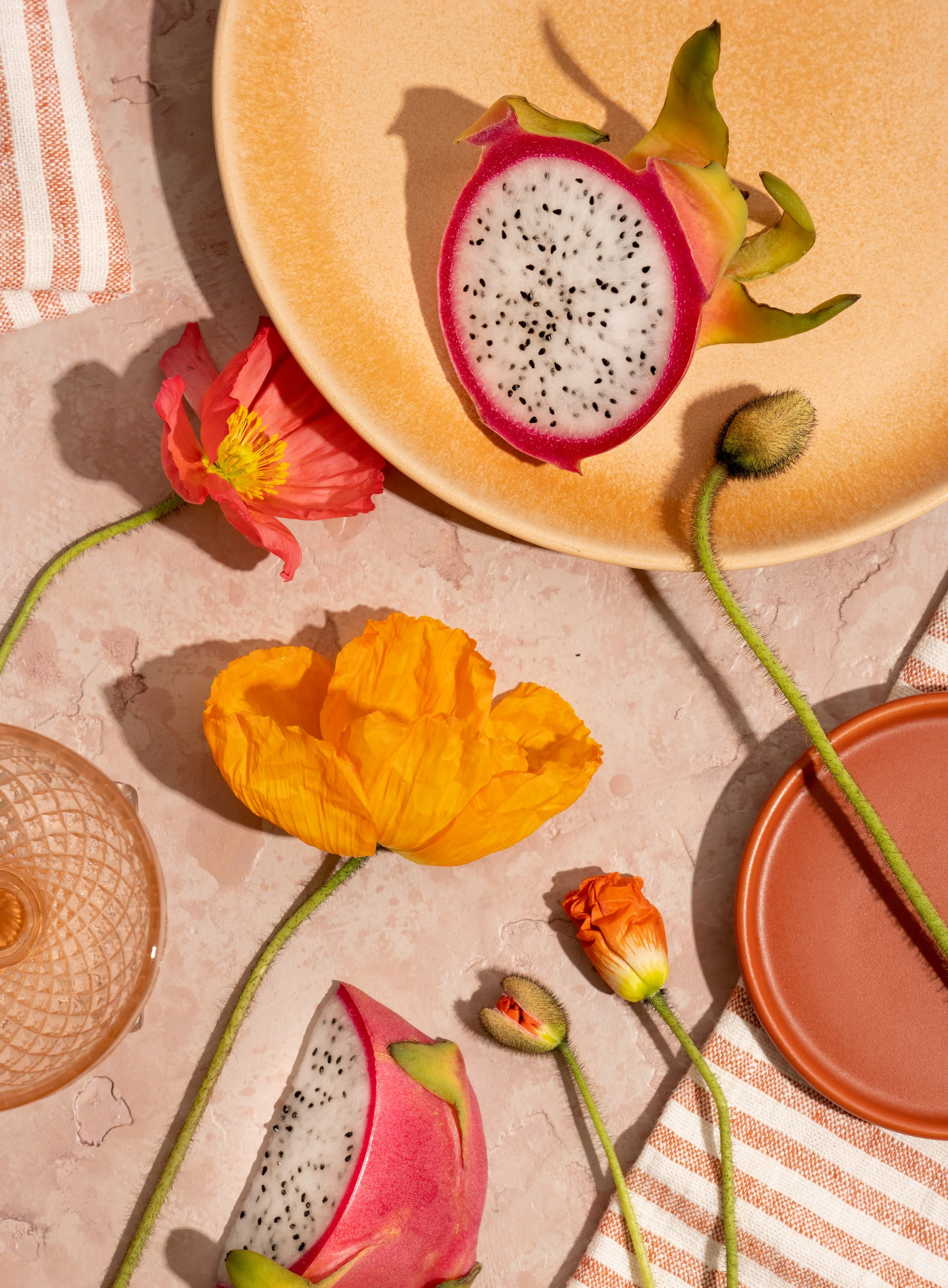 Half-cut dragon fruits, pink poppies, orange flowers, and a peach-colored plate with a striped napkin on a textured beige surface.
