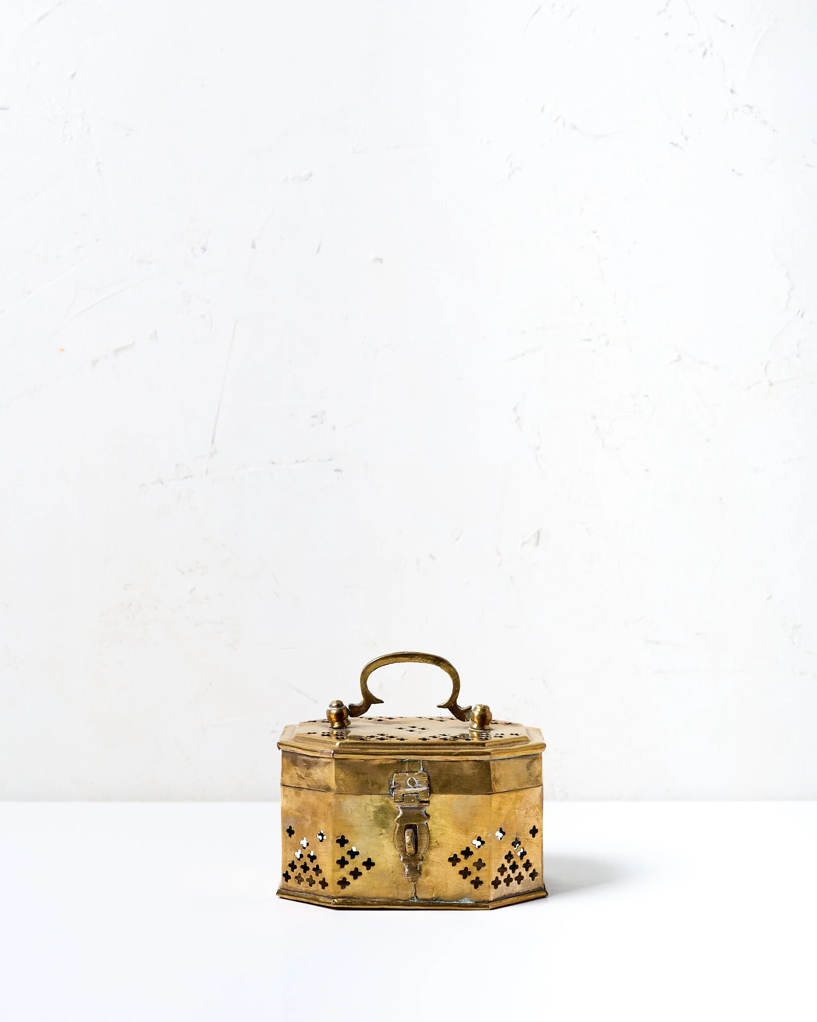 An antique small brass box with a handle, latch, and perforated sides, placed on a white surface against a white textured wall.
