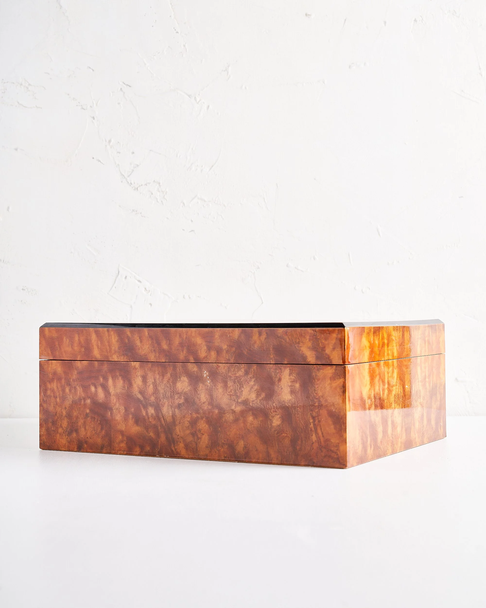A rectangular wooden cigar box with a polished surface and a natural wood grain pattern, placed on a white surface against a white textured wall.
