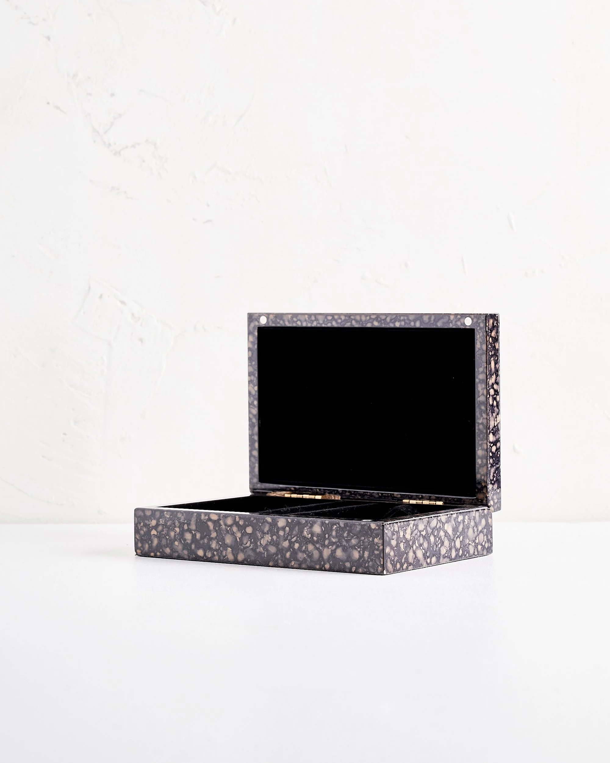 An open shallow rectangular jewelry box with a black and beige speckled pattern on white surface against a textured white wall.